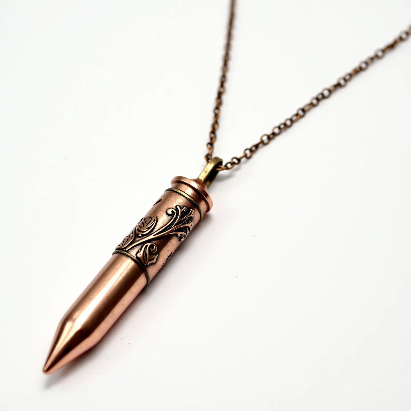 This bullet necklace features a metallic pendant in the shape of a bullet, crafted from what appears to be copper or a copper-colored material, exhibiting a warm, earthy hue. The pendant is intricately engraved with elegant designs, adding a decorative element to its otherwise sleek form. The chain, matching in tone, is formed of small, round links that provide a subtle, yet durable attachment for the pendant. The necklace is equipped with a small loop connector at the top of the bullet pendant to attach it securely to the chain. There are no visible gems adorning the necklace, maintaining a minimalist and rugged aesthetic.