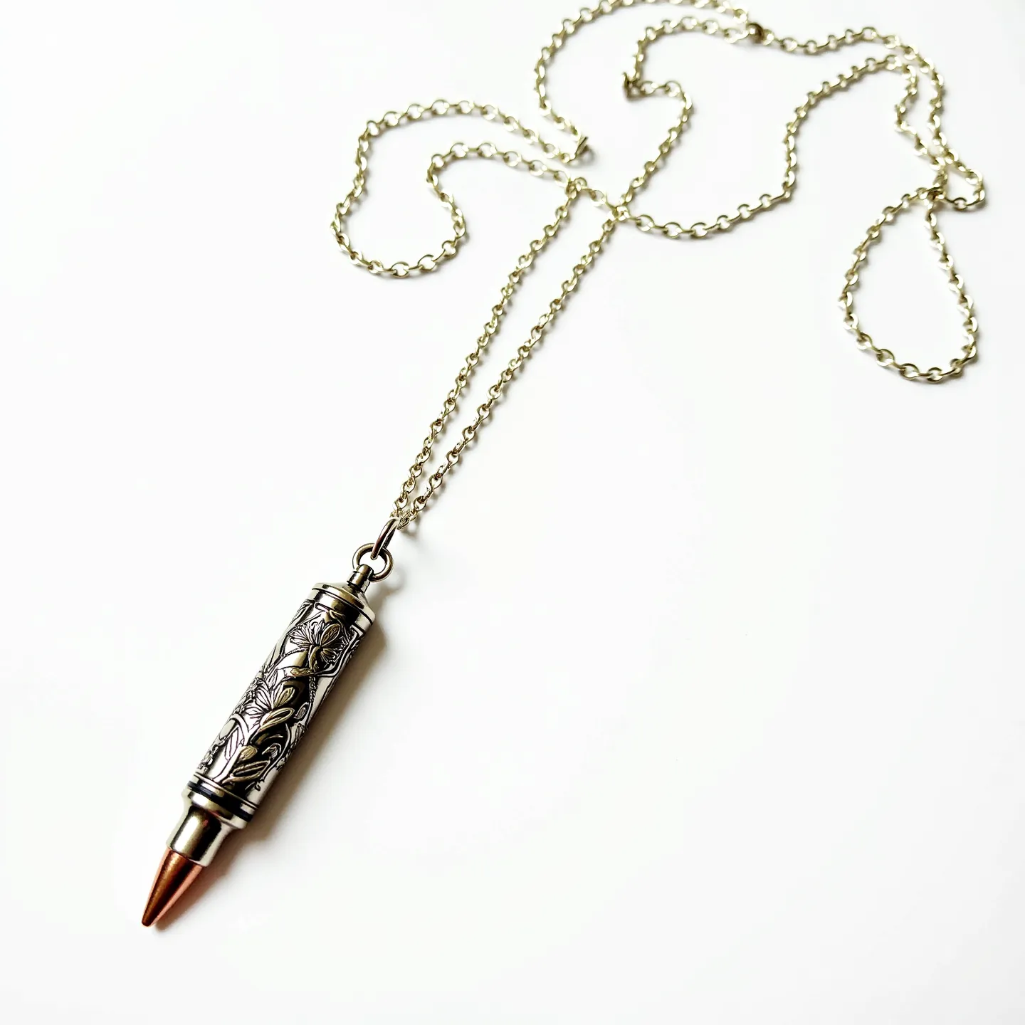 This bullet necklace features a metallic bullet pendant with an intricately engraved design, showcasing a floral pattern along the casing. The pendant appears to be crafted from a silvery metal, possibly stainless steel or another durable metal, while the tip is copper-toned, suggesting a two-tone design. The necklace is suspended from a delicate chain with a simple loop attachment at the top of the bullet, allowing the pendant to hang freely. The chain itself features small, uniformly sized links, providing a subtle yet elegant appearance.