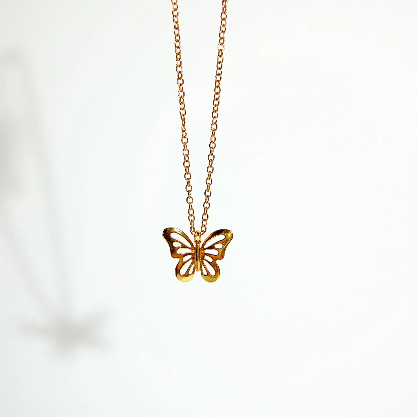 This butterfly necklace features a delicate butterfly pendant crafted from a smooth, metallic material, most likely gold or gold-plated, providing a warm and shiny appearance. The butterfly is intricately designed with cut-out spaces that lend an airy, lace-like quality to its wings. It is suspended from a fine chain composed of interconnected, oval links, maintaining a consistent metallic tone. The necklace is likely finished with a simple clasp mechanism, such as a lobster or spring ring clasp, to ensure ease of wear and secure attachment. The elegant design highlights both simplicity and sophistication.