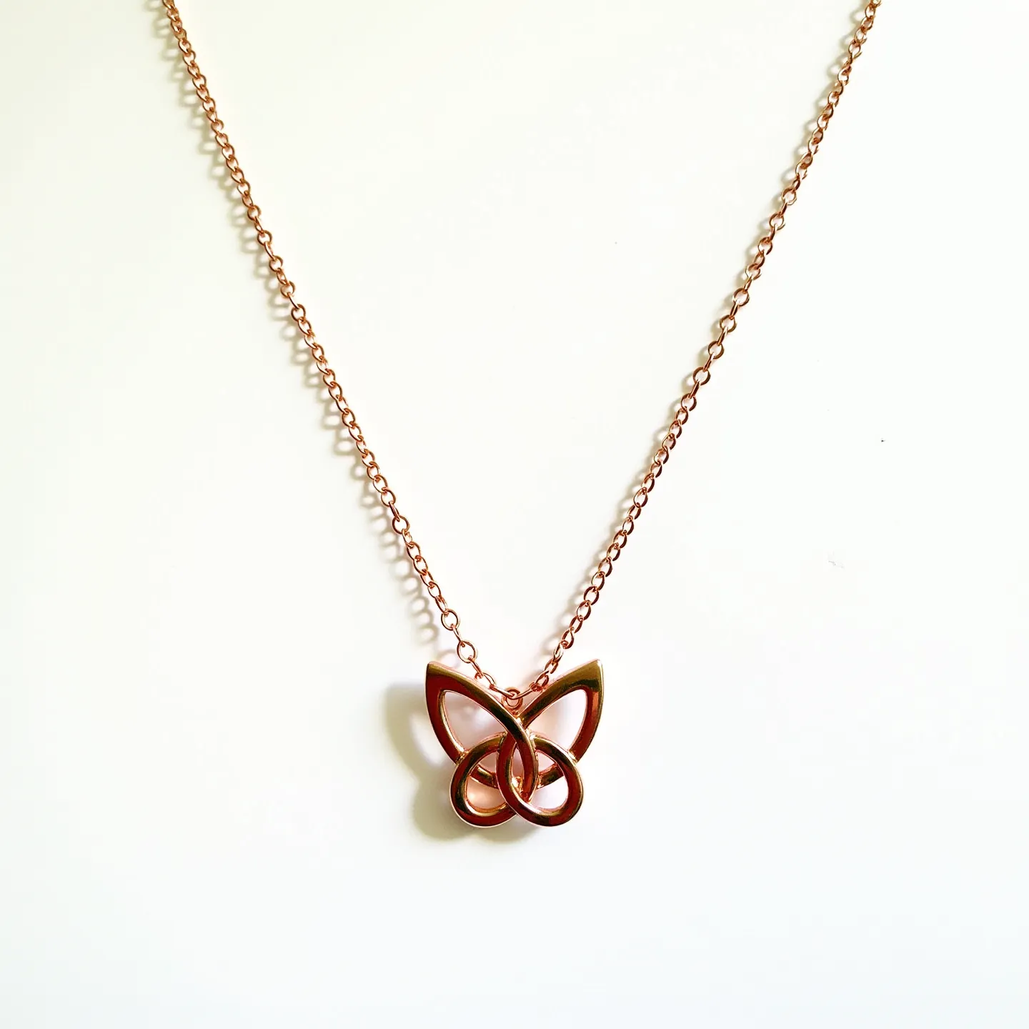 This butterfly necklace features a delicate, rose gold-colored metal pendant in the shape of a butterfly, characterized by smooth, intertwined loops forming abstract wings. The design exudes elegance with its streamlined contours and polished finish. The pendant is suspended from a matching fine chain, which complements the polished appearance of the butterfly. The necklace likely includes a typical lobster clasp for secure attachment, reflecting a balance between beauty and practicality. The overall design is modern and minimalist, making it suitable for a variety of occasions.