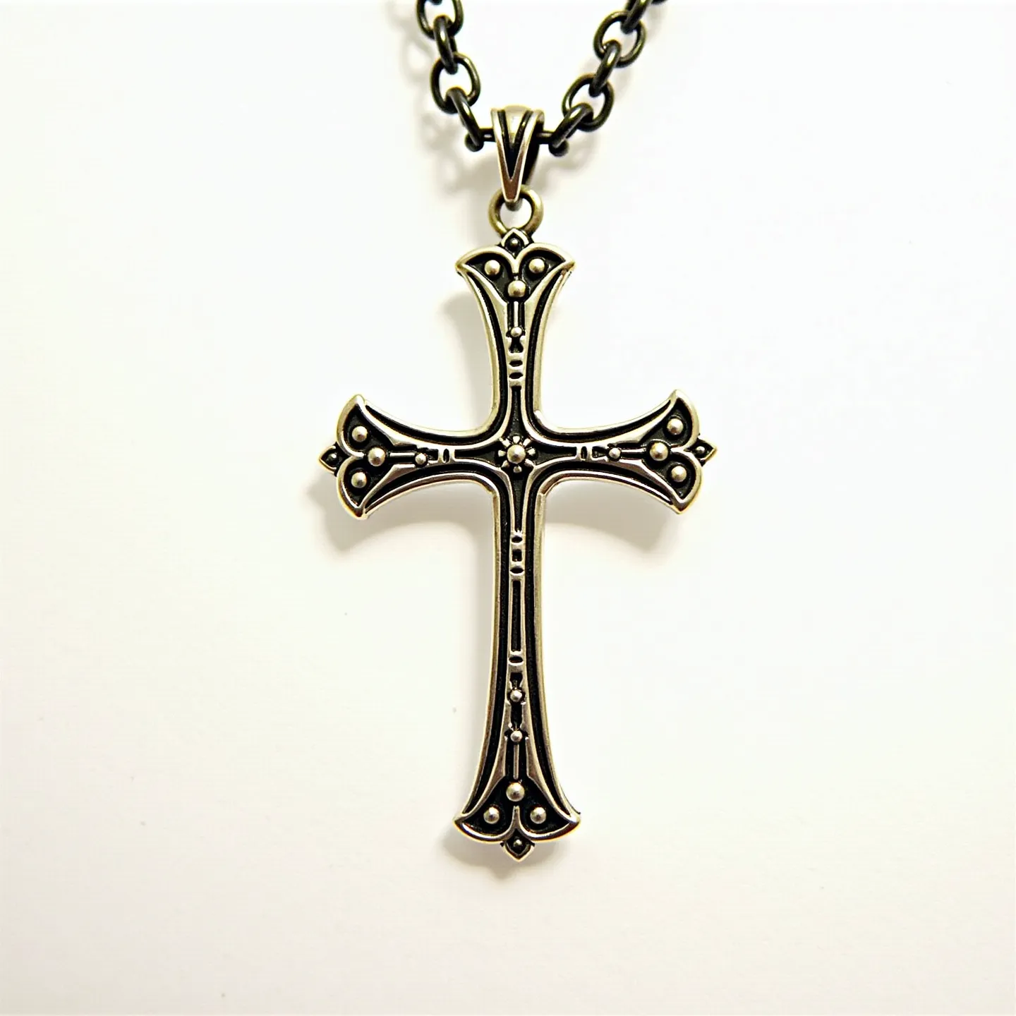 This catholic necklace features a striking and ornate cross pendant made of metal with an antique finish, showcasing intricate detailing along its arms. The pendant includes small circular embellishments resembling beads, strategically placed within the cross design to add texture and visual interest. It hangs from a robust, dark-toned chain with rounded links, providing a stark contrast to the lighter hue of the cross. The attachment to the chain is facilitated by a metal loop, securely holding the pendant. The overall aesthetic is both elegant and classic, reflecting traditional Catholic motifs.