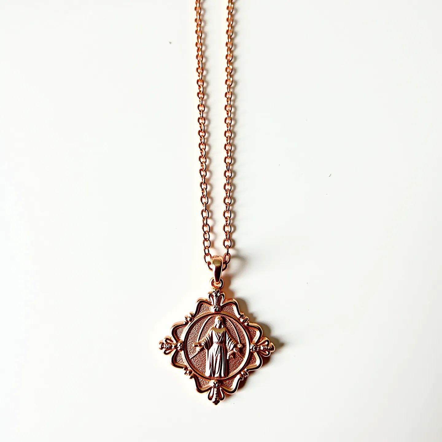 This catholic necklace features a rose gold colored medal pendant intricately designed with a religious figure, showcasing an ornate scalloped edge with decorative flourishes. The necklace includes a delicate chain crafted from what appears to be a matching metal, enhancing the overall elegance and unity of the piece. The pendant is smoothly integrated into the chain through a secure and coordinating looped bail. The chain structure appears to be of a standard link design, which complements the sophistication and simplicity of the pendant.