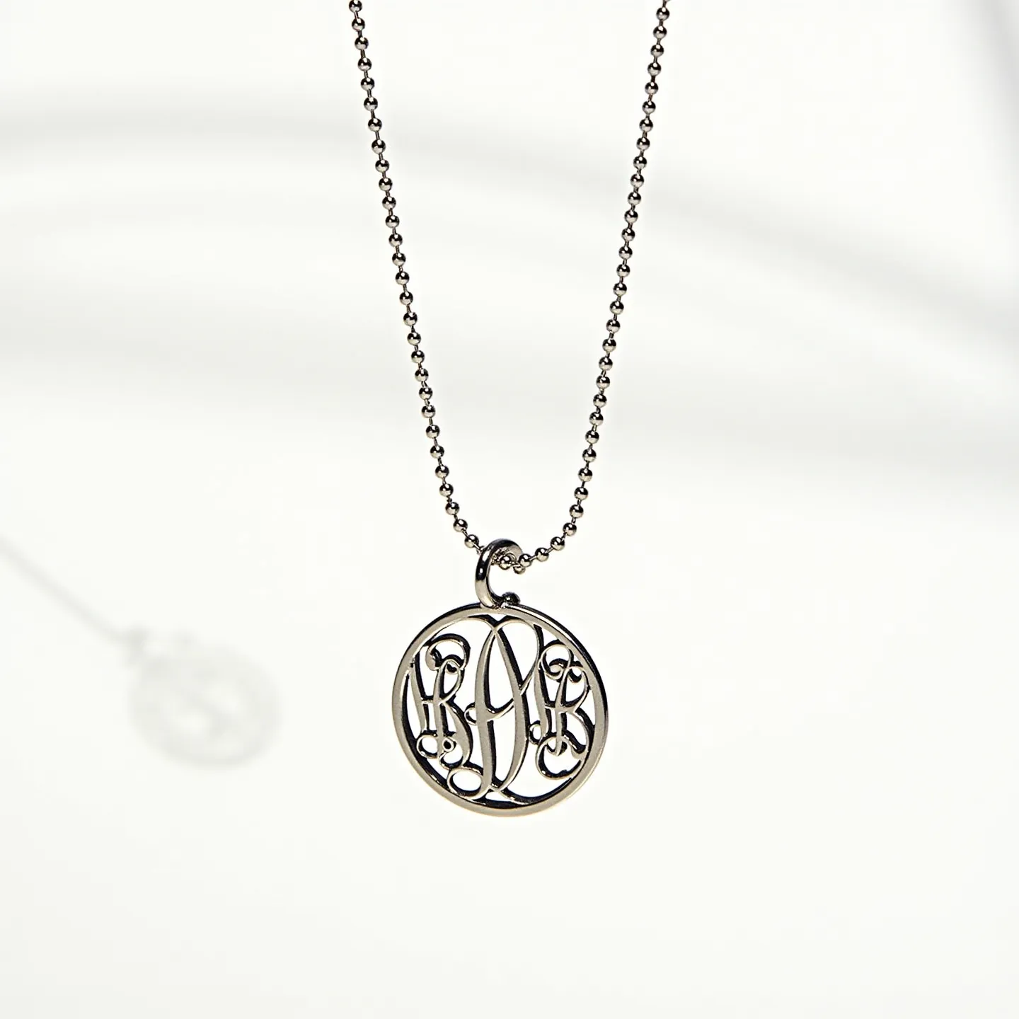 This charm necklace features a delicate round pendant with an intricate monogram design, crafted from a shiny metal that appears to be silver or white gold. The pendant is suspended from a fine ball chain, indicating a light and simple construction. The necklace includes a standard loop for the attachment, making it easy to open and close with a small clasp. The design of the monogram within the pendant provides a touch of personalization, showcasing elegant curves and loops that form the letters in a sophisticated style.