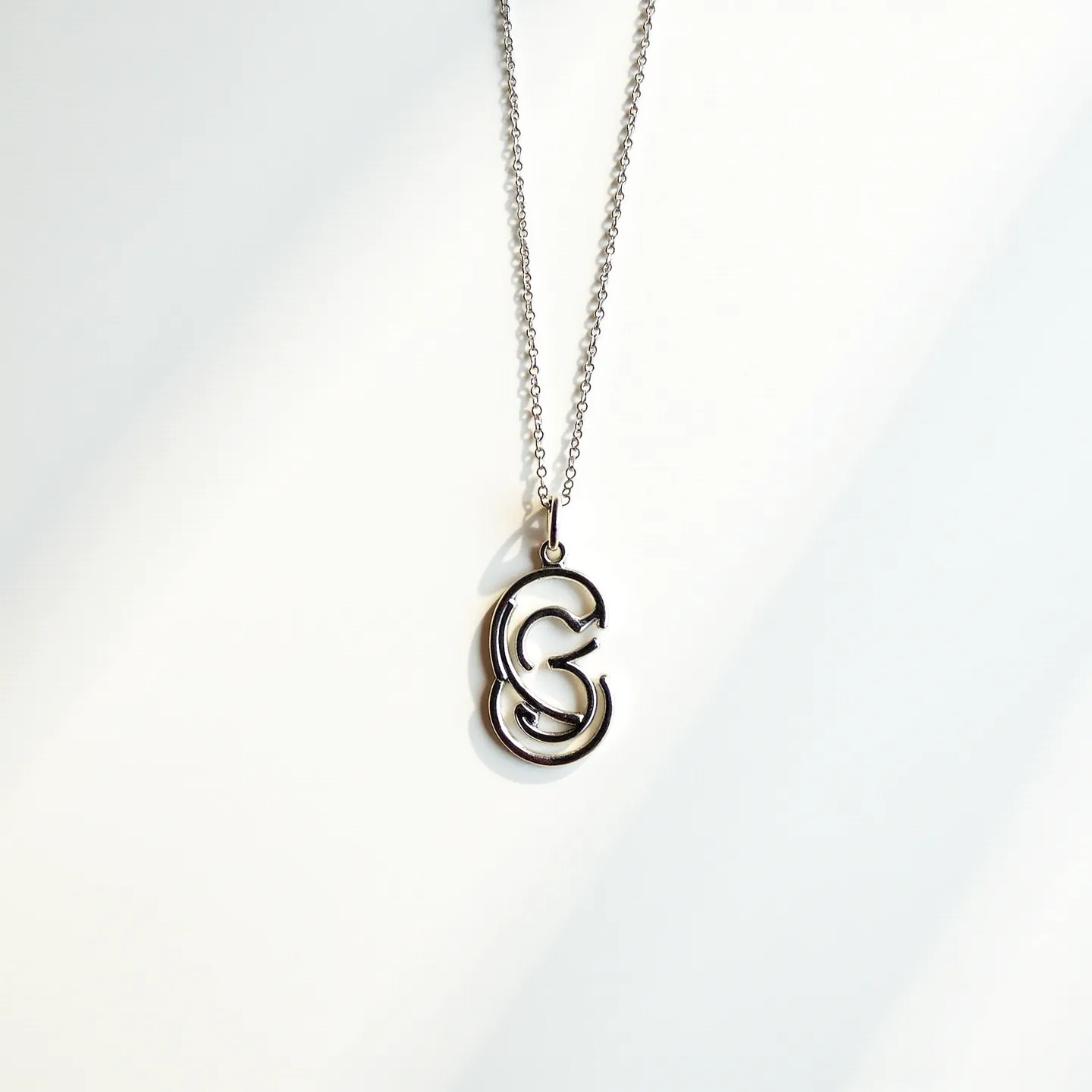 This charm necklace features a delicate chain made of a metallic material, likely stainless steel or silver, with a lustrous finish that catches the light. The centerpiece is a stylish charm in the shape of an abstract or stylized 'G', crafted from the same metallic material as the chain, ensuring a cohesive look. The charm is suspended from the chain by a small metal loop that connects to a simple yet secure clasp mechanism. The minimalist and modern design of the charm, along with the fine detailing of both the chain and pendant, lends a contemporary elegance to the piece.