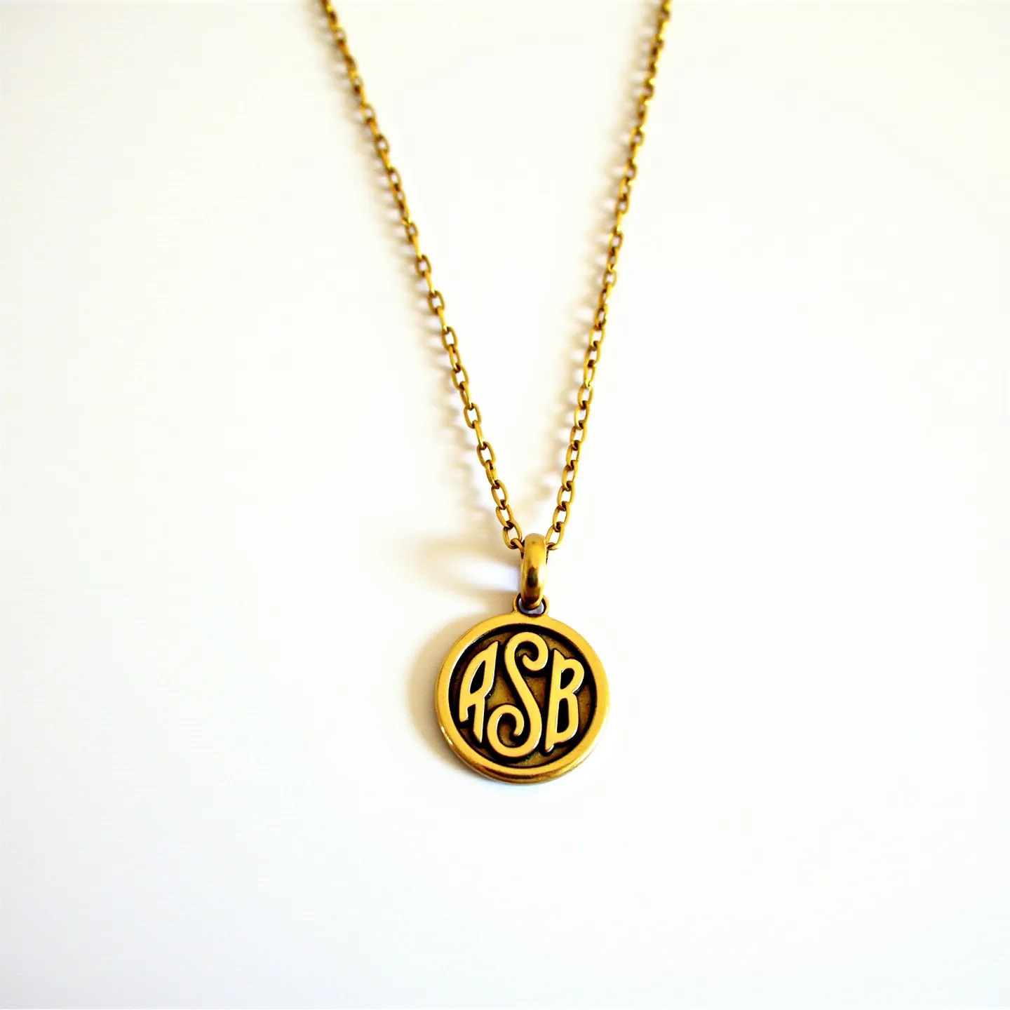 This charm necklace features a finely crafted round pendant with an engraved monogram design, finished in a warm gold tone. The necklace is composed of a delicate, yet sturdy, chain that matches the pendant's coloring, suggesting a metal like gold or a gold-toned alloy. The pendant hangs from a simple loop attachment that seamlessly integrates with the chain's design. The overall aesthetic of the necklace is elegant, with the monogram adding a personalized touch.