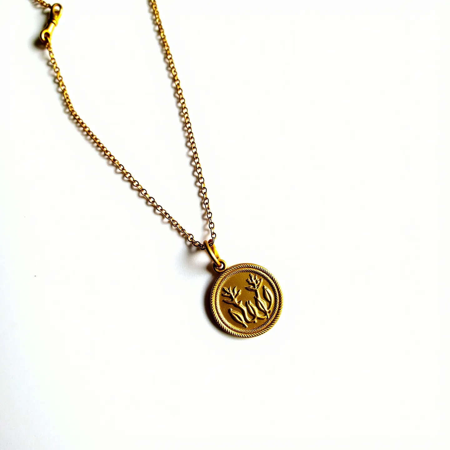 This charm necklace features a delicate gold chain with an intricately designed pendant. The pendant is circular with a raised floral design, framed by a textured border. The material appears to be gold-toned metal, lending an elegant and timeless appeal to the piece. It is attached to the chain with a simple loop bail, ensuring it hangs elegantly. The chain is secured with a lobster clasp, providing a sturdy and reliable closure.