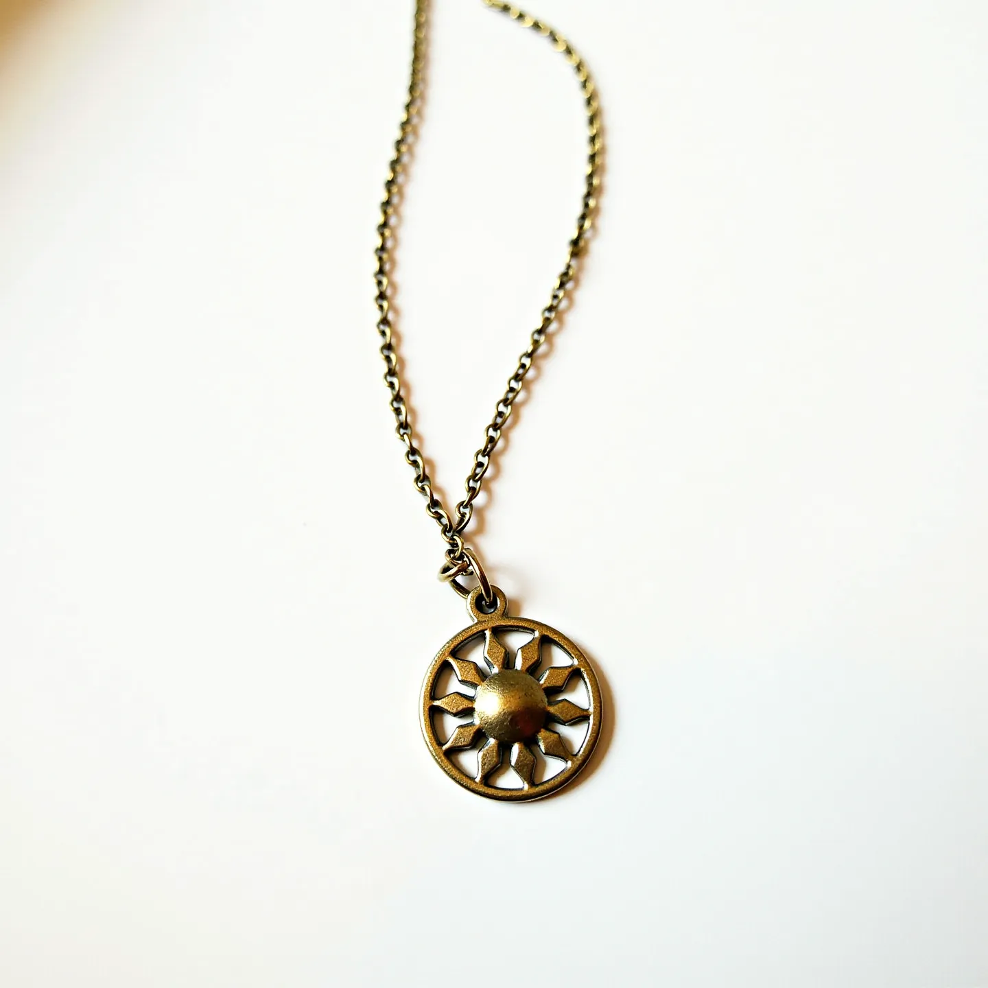 This charm necklace chain features a delicate link chain made of a bronze or brass-like metal. The pendant is a circular charm with a sun motif, showcasing a central raised dome surrounded by sun rays set in an openwork design. The charm adds an artistic and symbolic element to the piece. The necklace is equipped with a standard clasp, ensuring secure and convenient wearability.