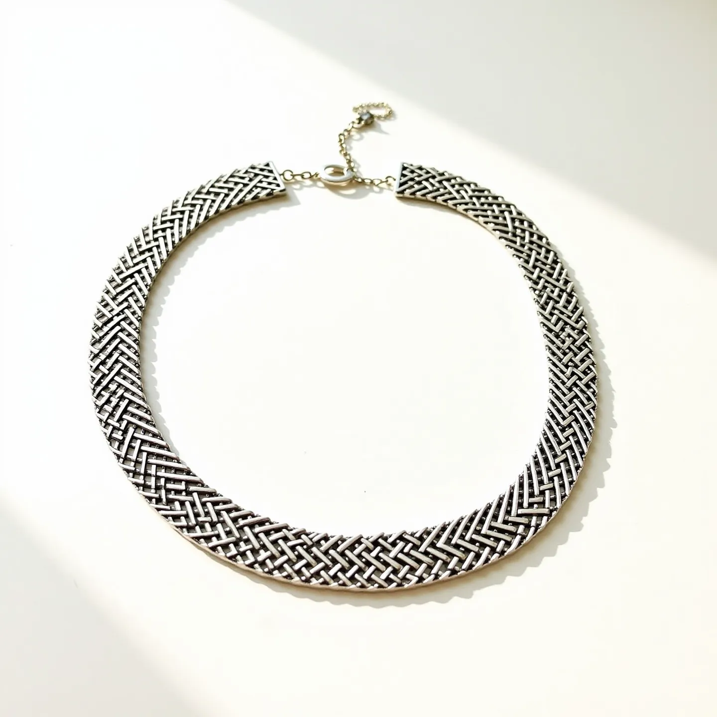 This choker chain features an intricately woven design, with interlaced metal strands creating a textured, braided appearance. The material appears to be a polished metal, likely silver or a silver-toned alloy, giving it a sleek and shiny finish. The chain is complemented by a lobster clasp, which is both functional and secure, allowing for easy adjustment and closure. The overall design evokes a sense of elegance and sophistication, making it a versatile accessory for various occasions.