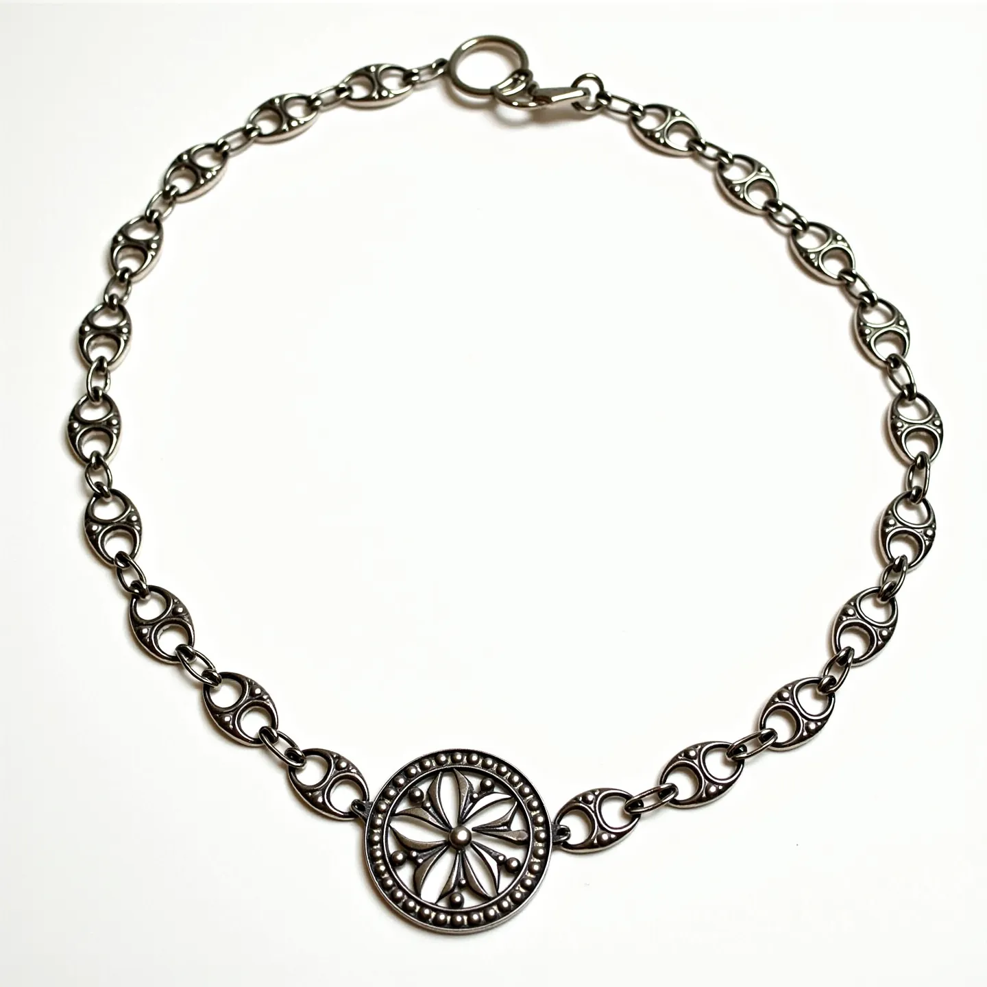 This choker chain features a series of intricately designed metal links, each formed in a stylized openwork pattern that gives a distinct, cohesive appearance. At its center, a round pendant with a decorative, flower-like motif takes prominence, bordered by small beaded detailing. Made of a silvery metal, the choker showcases a polished finish that enhances its aesthetic appeal. The use of intricate cutwork in the central pendant adds an element of craftsmanship. The choker is secured with a straightforward ring clasp, ensuring easy wearability while maintaining the overall sleek design.