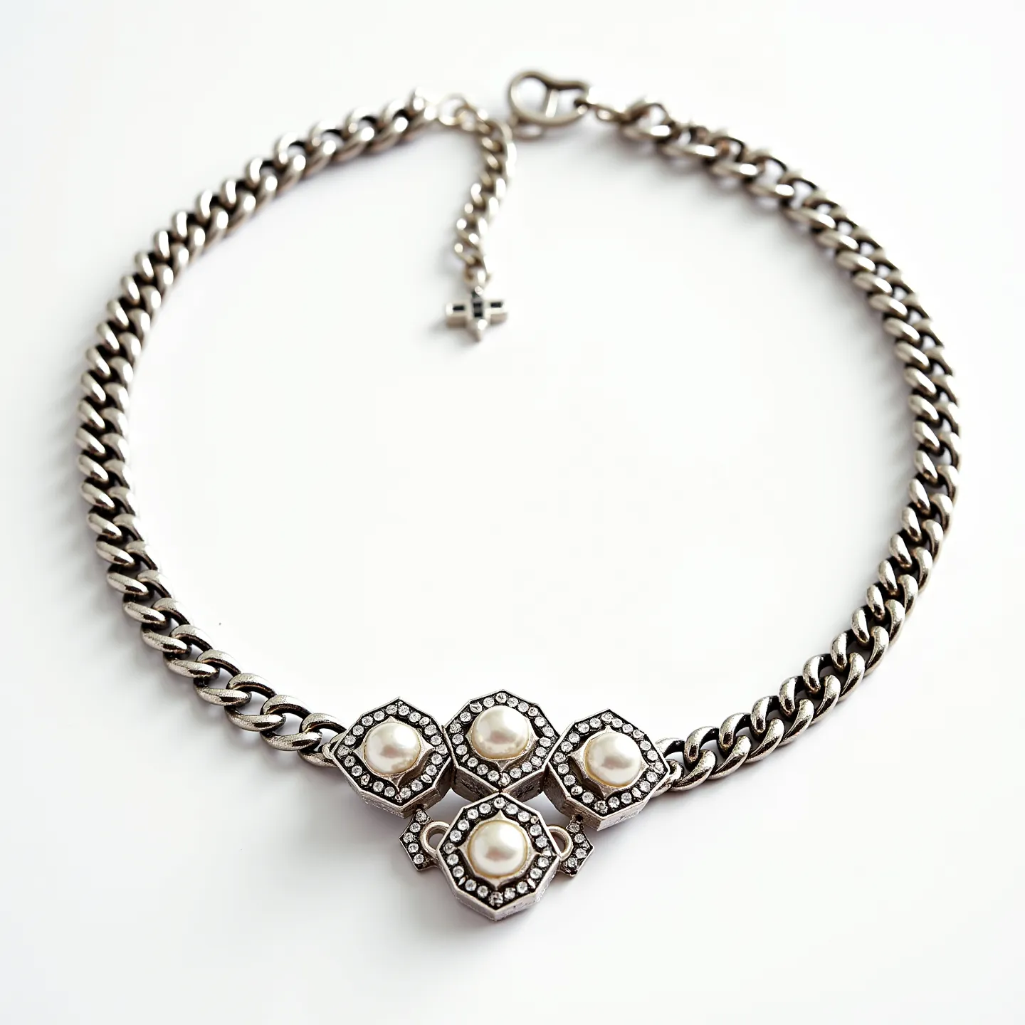 This choker chain features a robust, metallic-link design, likely crafted from a durable metal such as stainless steel or silver. At its center, it showcases a striking arrangement of four hexagonal settings, each adorned with a rounded pearl as the central gem. The pearls are surrounded by a series of smaller, clear stones, possibly diamonds or crystals, adding a touch of elegance and sparkle. The settings are positioned in a symmetrical pattern, creating a visually appealing focal point. The choker is secured with a sturdy clasp, accompanied by an adjustable chain that ends in a small cross-shaped charm, providing both functionality and style to the piece.