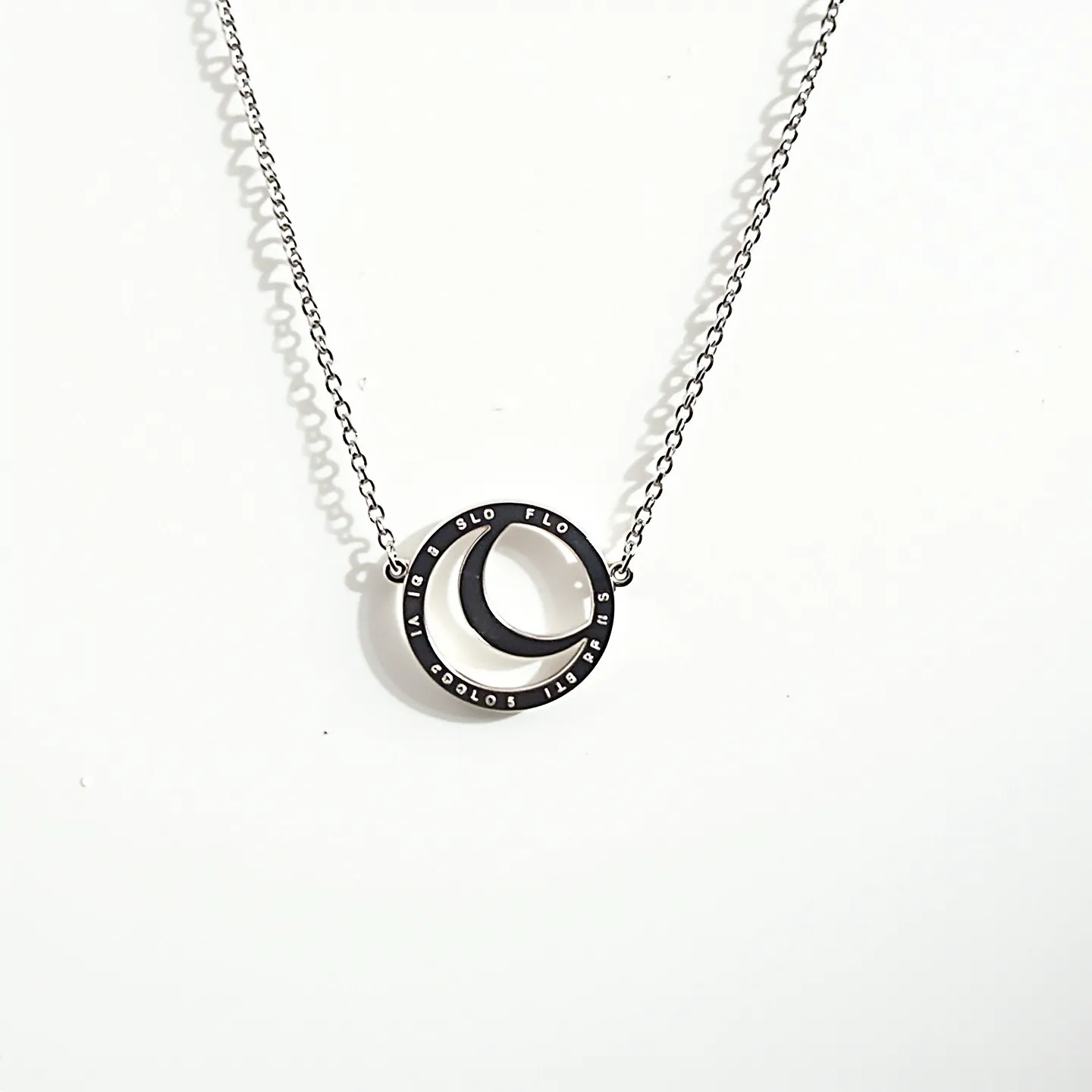 This circle necklace features a contemporary design with intertwined circular pendants. The primary pendant is a smooth metal circle, possibly stainless steel or sterling silver, with a secondary crescent-shaped element inside. The surface of the outer circle has engraved text, adding a unique and modern touch. The necklace is suspended from a delicate metal chain that complements the minimalist design. The chain appears to be fastened with a standard clasp, ensuring secure and easy wearability. The overall style is sleek and versatile, suitable for various occasions.