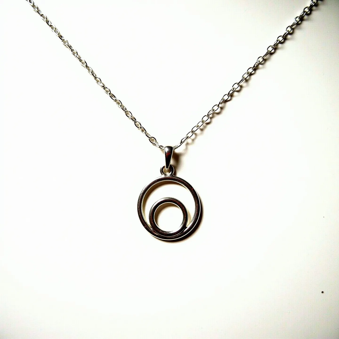 This circle necklace features a minimalist design with two concentric circular pendants made of a shiny metallic material, likely stainless steel or silver. The pendant is suspended from a delicate chain composed of small, evenly spaced links, offering a subtle yet elegant appearance. A small, smooth bail securely attaches the pendant to the chain, allowing it to hang neatly. The necklace’s simple design focuses on the smooth curves and reflective surface of the metal, without any gemstones or additional embellishments. A typical spring ring clasp or lobster clasp is likely used for fastening, providing both security and ease of wear.