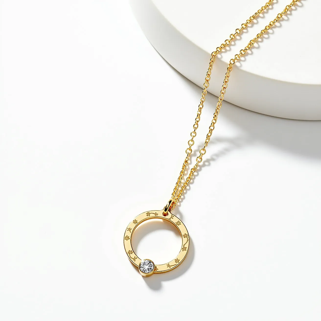 This circle necklace features a gold-toned circular pendant adorned with a pattern of small star engravings, adding a delicate decorative element to its design. Attached to the bottom of the circle is a round, brilliant-cut gemstone set in a bezel setting, which offers both security and a sleek look. The pendant is suspended from a fine chain link necklace, which complements the pendant with its matching gold tone. The necklace includes a simple and functional clasp, allowing for easy wear and secure attachment.
