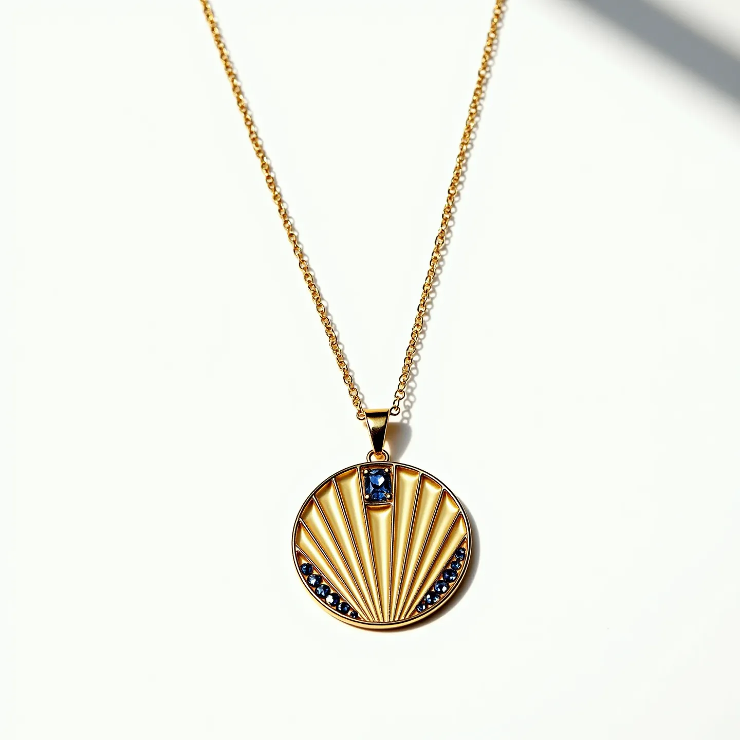 This circle necklace features a gold pendant with a geometric design resembling a stylized shell or fan. At the top center of the pendant is a rectangular blue gemstone, which appears to be a faceted sapphire or similar stone, set in a bezel setting. Below, the design is accentuated with additional smaller round blue stones that are prong-set along the curved bottom edge of the pendant. The necklace is suspended from a delicate gold chain, which is likely made of the same material as the pendant, providing a cohesive and elegant look. The chain appears to have an integrated attachment that seamlessly connects to the pendant, offering a polished finish.