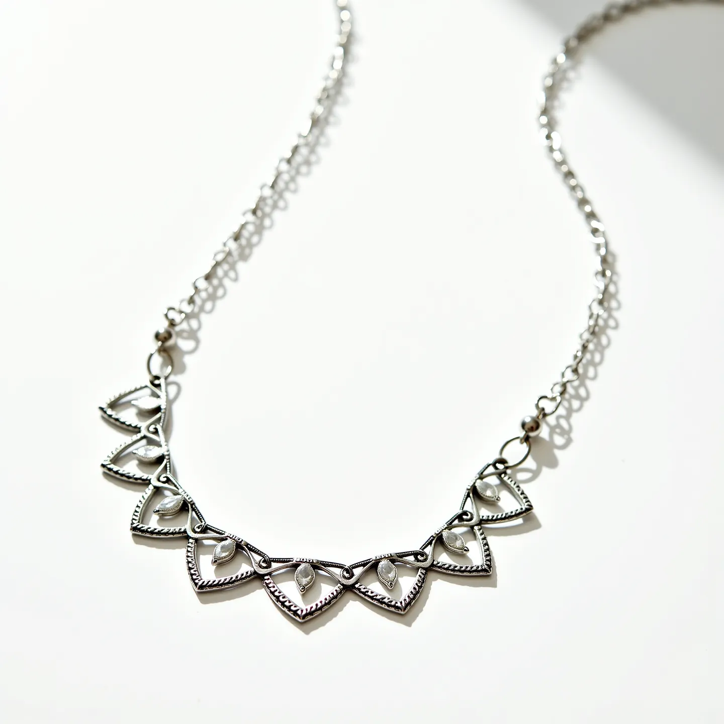 This circle necklace features intricate metalwork with a series of triangular elements linked together to form the central design. It appears to be made from a silver-toned metal, possibly sterling silver, giving it a sleek and polished finish. Each triangular segment is adorned with a small, teardrop-shaped embellishment, adding a touch of elegance to the design. The necklace is connected by a delicate chain with interlocking links that complement the pendant's motif. It fastens with a simple jump ring, providing secure closure and ease of wear while maintaining the overall aesthetic appeal.