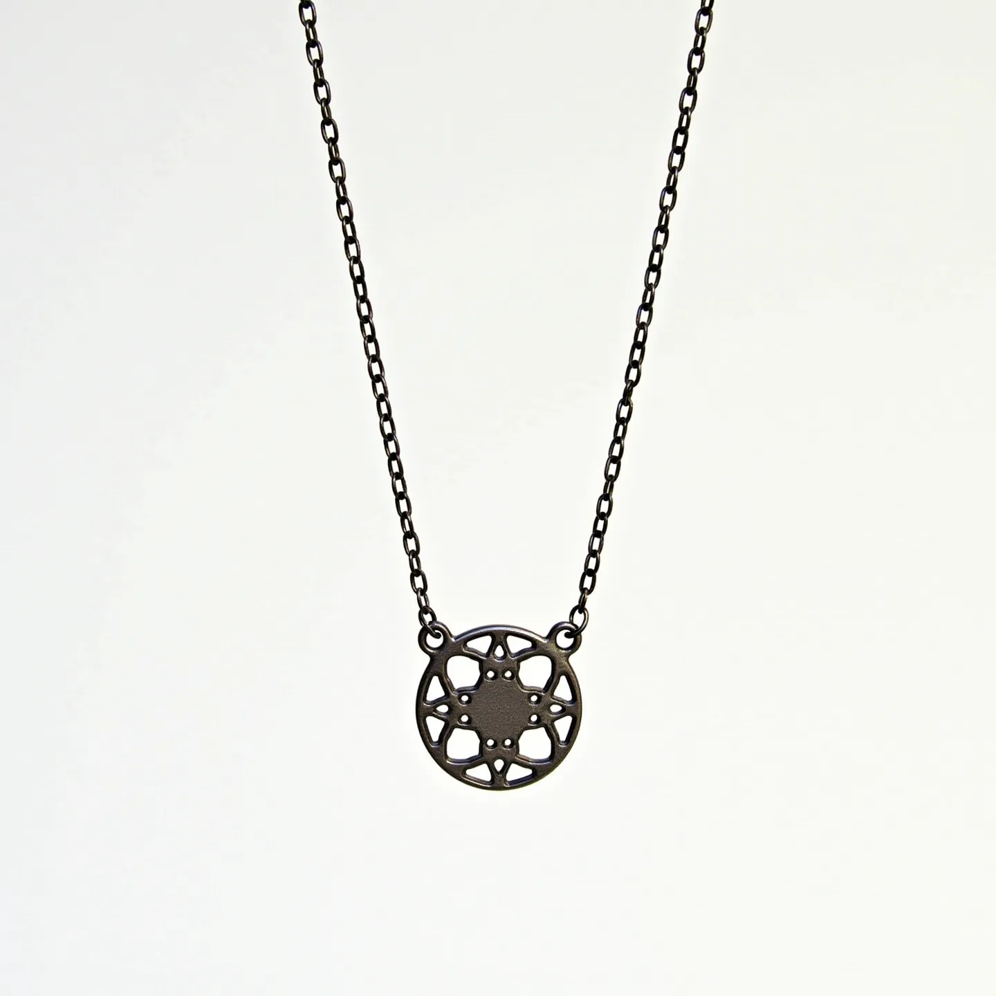 This circle necklace features an intricately designed pendant, made from what appears to be a metal with a dark matte finish. The pendant showcases a circular pattern that resembles a geometric or floral motif, centered around a star-like shape. The chain is composed of small, uniformly shaped links, suggesting a strong yet delicate construction that complements the pendant's intricate design. The necklace's clasp, a classic lobster clasp, provides a secure and adjustable attachment, making it versatile for various wearers.