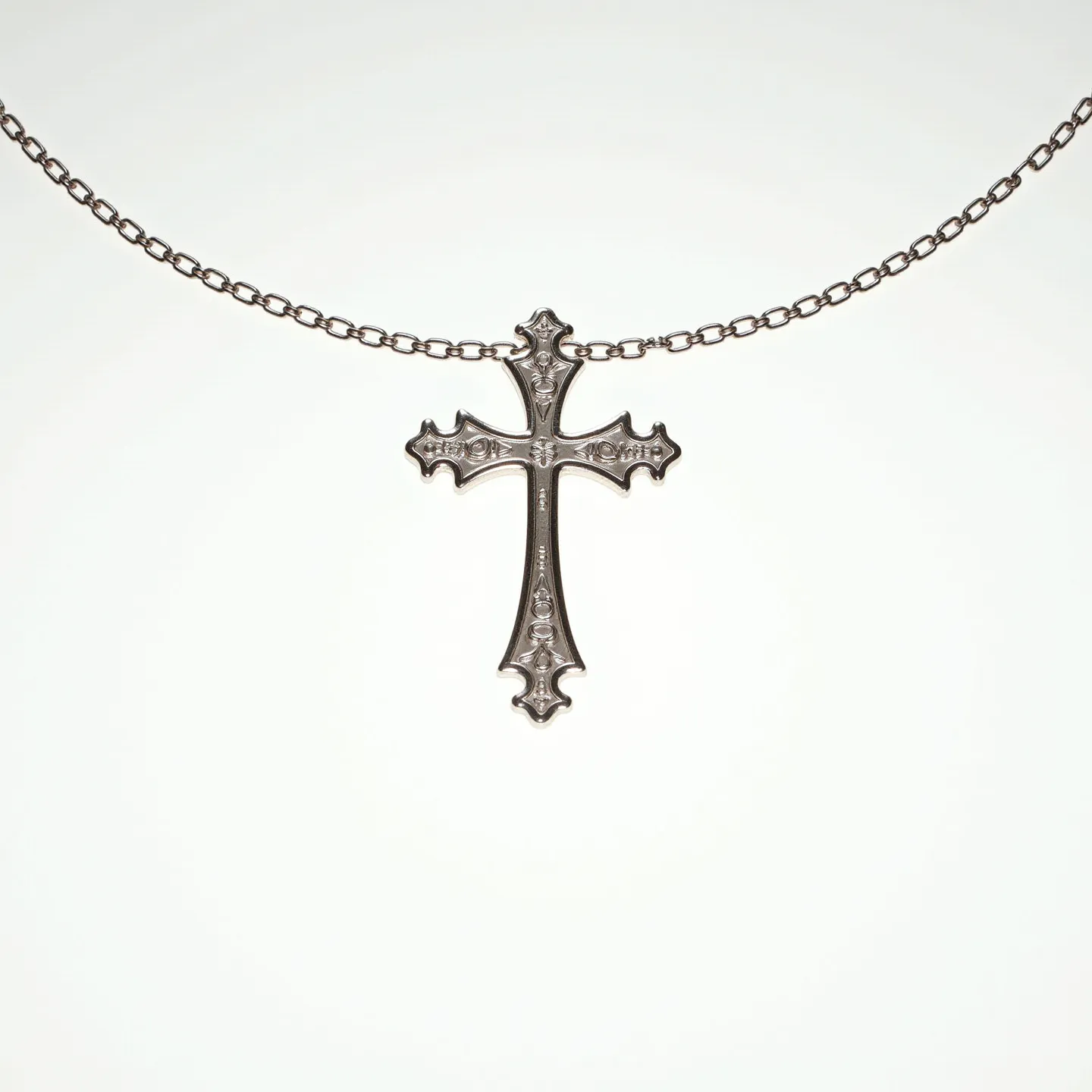 This cross necklace features a finely crafted metallic cross pendant with intricate detailing along its surface, evoking a classic, ornate appearance. The material appears to be a polished metal, possibly silver or stainless steel, which gives it a lustrous finish. The chain is a simple, yet elegant link chain that complements the pendant without overpowering it. The cross does not appear to have any gemstones embedded but is distinguished by its stylized floral-like extensions at each end, adding to its decorative allure. The necklace is likely secured with a standard clasp, ensuring ease of wearability while maintaining a cohesive, stylish design.