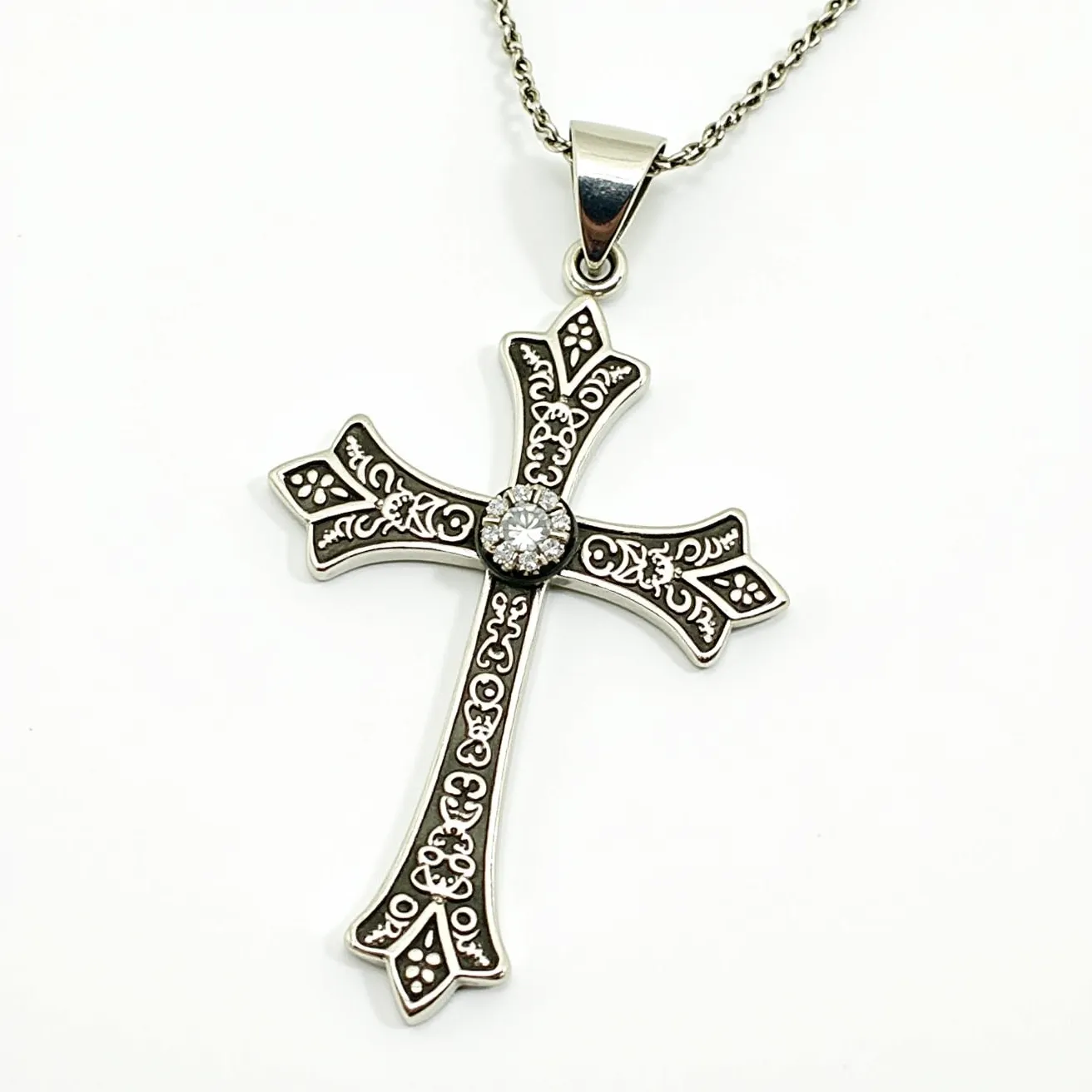 This cross necklace features an intricately designed metal cross with an ornate pattern, possibly made of silver or a silver-toned material. At the center of the cross, there is a round-cut gemstone, likely a diamond or crystal, set in a bezel or halo setting that enhances its brilliance. The cross is attached to a delicate chain via a classic bail clasp, allowing for secure wear. The detailed craftsmanship on the cross highlights elegant scrollwork and floral motifs, adding a touch of sophistication and timeless elegance to the piece.