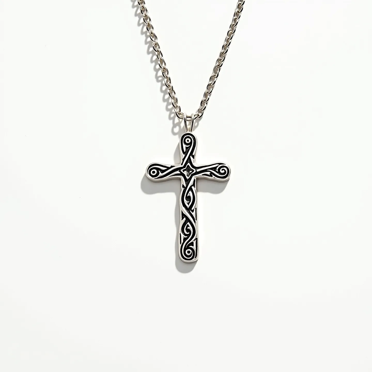 This cross necklace features a detailed design with intricate patterns etched into a metallic surface, likely made of silver or a silver-toned material. The cross pendant showcases swirling, Celtic-inspired motifs that give it a unique and artistic appearance. The edges of the cross are smooth, complementing the detailed intricacies within. The pendant is attached to a sturdy chain via a simple bail, which seamlessly connects it to the necklace. The chain itself, composed of interlinked, polished metal loops, provides both durability and a complementary aesthetic to the ornate cross.