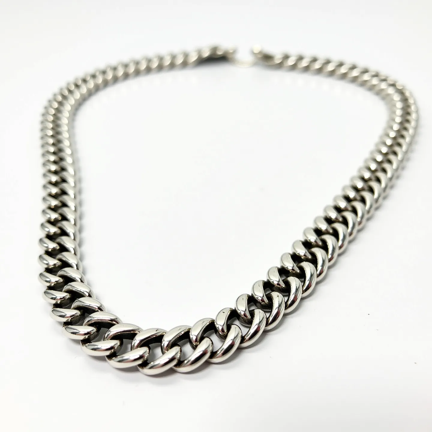 This curb chain features a classic, interlocking pattern of flat, oval links, giving it a sleek and polished appearance. The material appears to be a shiny metal, possibly stainless steel or silver, known for its durability and reflective surface. The chain does not exhibit any gemstones or additional ornamentation, focusing instead on the simple elegance of its tightly knit links. A lobster clasp is evident at the end, providing a secure and easy method of fastening. The chain's design and construction contribute to a stylish yet understated accessory, suitable for various occasions.