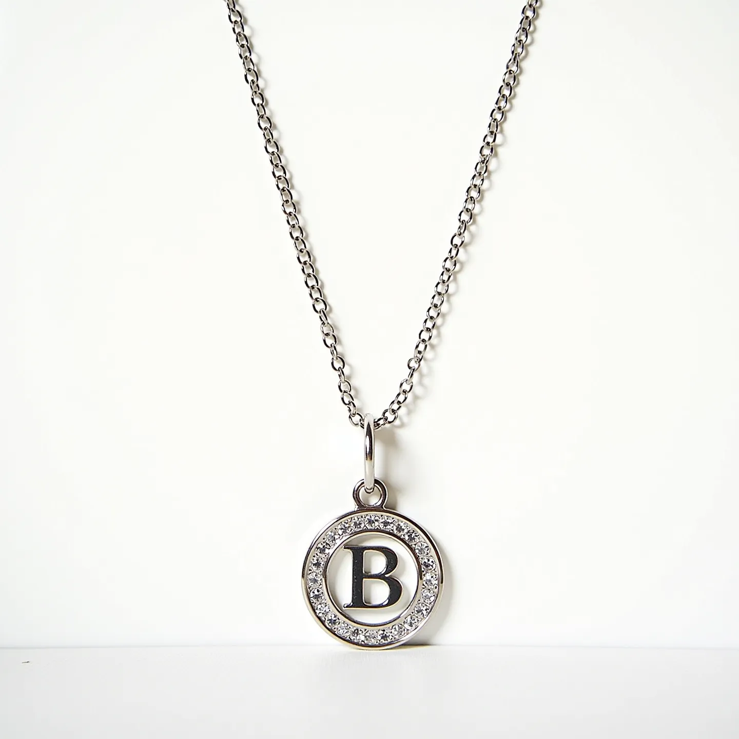 This custom charm necklace features a delicate chain, likely crafted from silver or white gold, which supports a circular pendant adorned with a prominent letter "B" in the center. The charm is encircled by a series of small, sparkling clear stones, which appear to be set in a pavé style, enhancing the necklace's elegant appeal. The attachment is a simple loop, seamlessly integrating the pendant with the chain, and providing a classic and minimalist aesthetic.