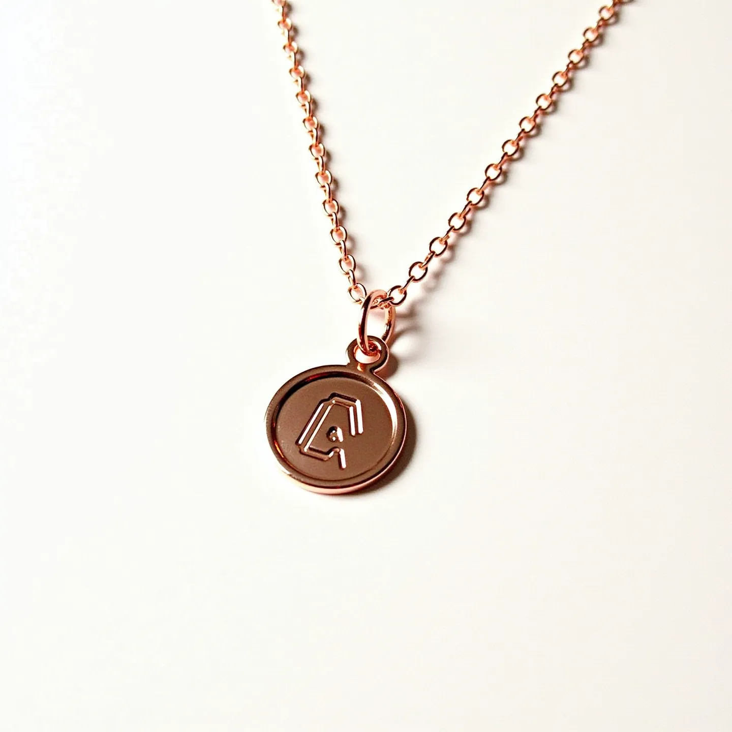 This custom charm necklace features a delicate rose gold chain with a round pendant. The pendant is crafted from a similar rose gold metal, which showcases an engraved letter "A" in a stylized font. The chain consists of interlinked round links that provide an elegant and cohesive appearance, complementing the minimalist aesthetic of the charm itself. A simple loop connects the charm to the chain, ensuring a secure attachment. The necklace is closed with a small, matching spring ring clasp, adding to its seamless design and functionality.