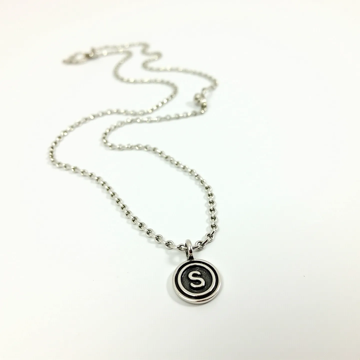 This custom charm necklace features a sleek, metallic chain composed of interlinked round beads, likely made from stainless steel or a similar durable metal, creating a polished and modern appearance. At its center hangs a circular charm engraved with the letter "S," framed in a contrasting hue that provides depth and visual interest. The charm is securely attached to the chain via a simple loop, which complements the overall minimalist design. The necklace is finished with a classic lobster clasp, ensuring a secure and adjustable fit for the wearer.