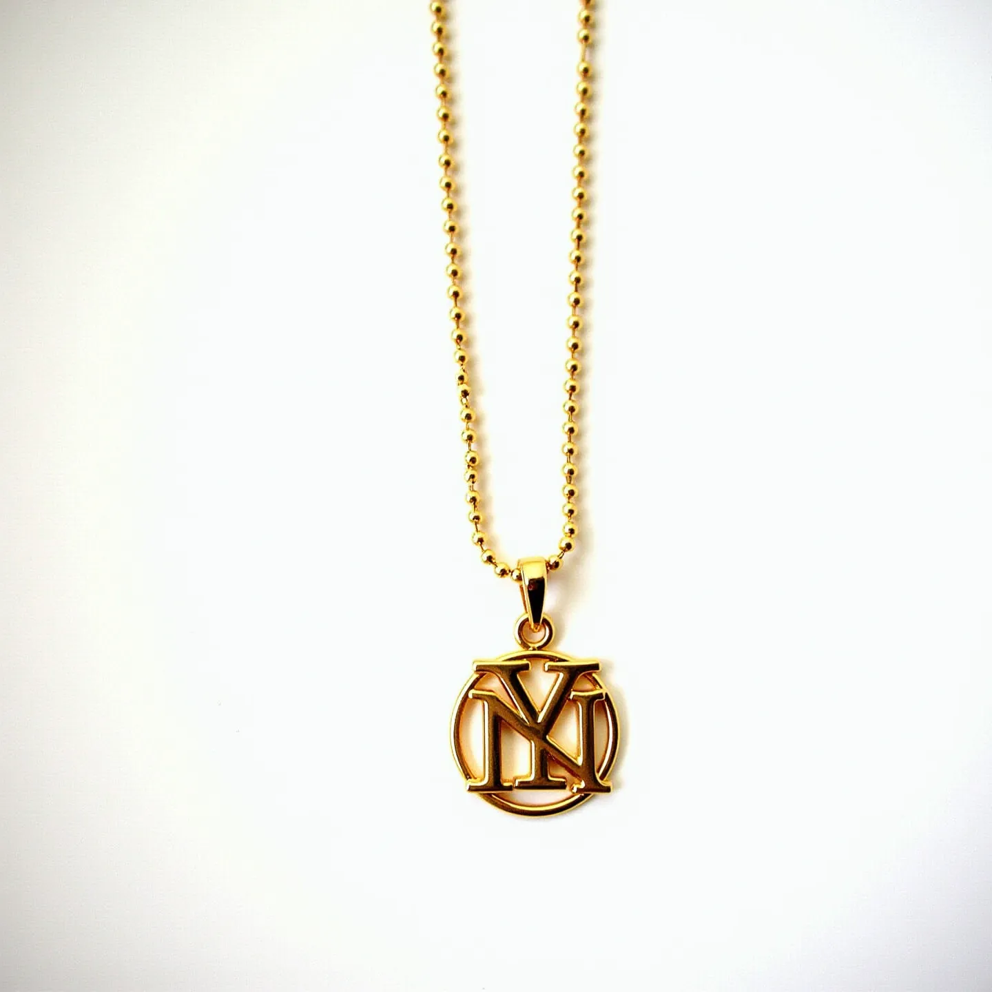 This custom charm necklace features a gold-tone metal chain composed of small, round beads, giving it a polished and elegant look. The pendant displays an intricate, cut-out design in the shape of stylized letters, also in a matching gold-tone. The charm hangs from a sleek, looped attachment that seamlessly connects to the chain, ensuring a cohesive design. The necklace is equipped with a standard clasp to secure it around the neck. The overall appearance of the necklace is stylish and personalized, with well-crafted metal elements.