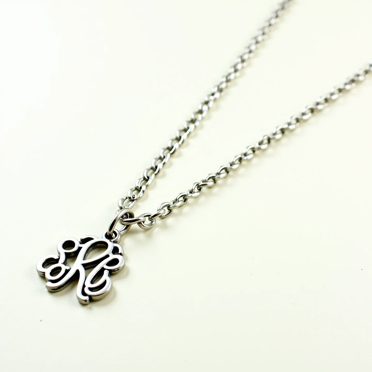 This custom necklace features a polished silver-tone metal chain with moderately sized links, creating a classic look. The center of the necklace showcases an intricate, script-style monogram charm, also in a matching silver-tone finish, adding a personalized touch to the piece. The pendant is connected to the chain through a simple, unobtrusive loop, allowing it to dangle freely. The necklace is likely secured with a standard lobster claw clasp, providing both security and ease of wear. Overall, the design emphasizes elegance with a personalized flair.