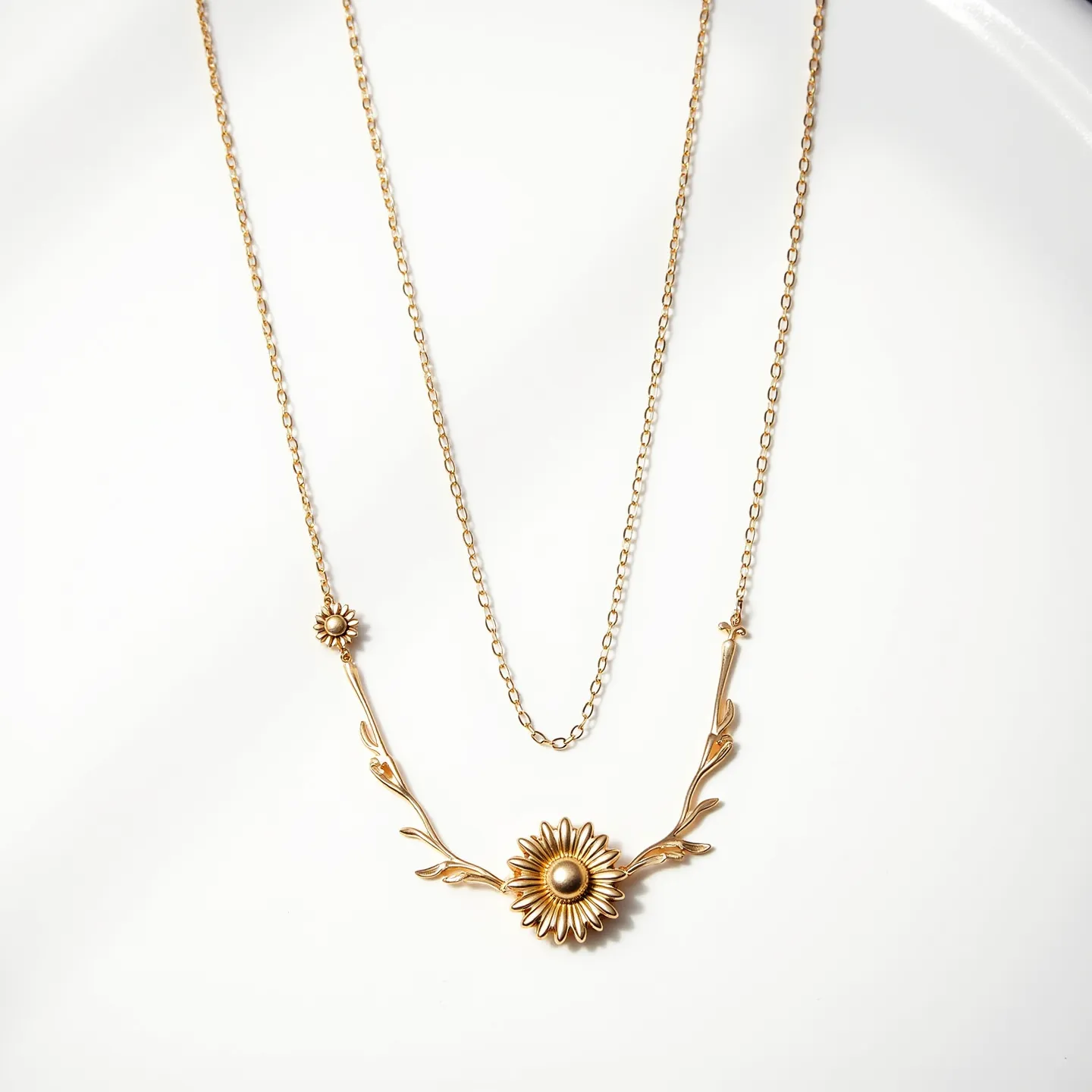 This daisy necklace features a delicate design with two daisy motifs crafted from a gold-toned metal. The central daisy is larger and intricately detailed, flanked by leaf-like embellishments that branch elegantly from the pendant’s main structure. The smaller daisy appears along one of the connecting chains, adding a charming asymmetrical detail. The necklace is composed of fine chain links that enhance its dainty aesthetic. It's likely secured with a standard clasp mechanism typical in necklaces of this style, ensuring ease of wear while complementing the overall lightweight and graceful design.