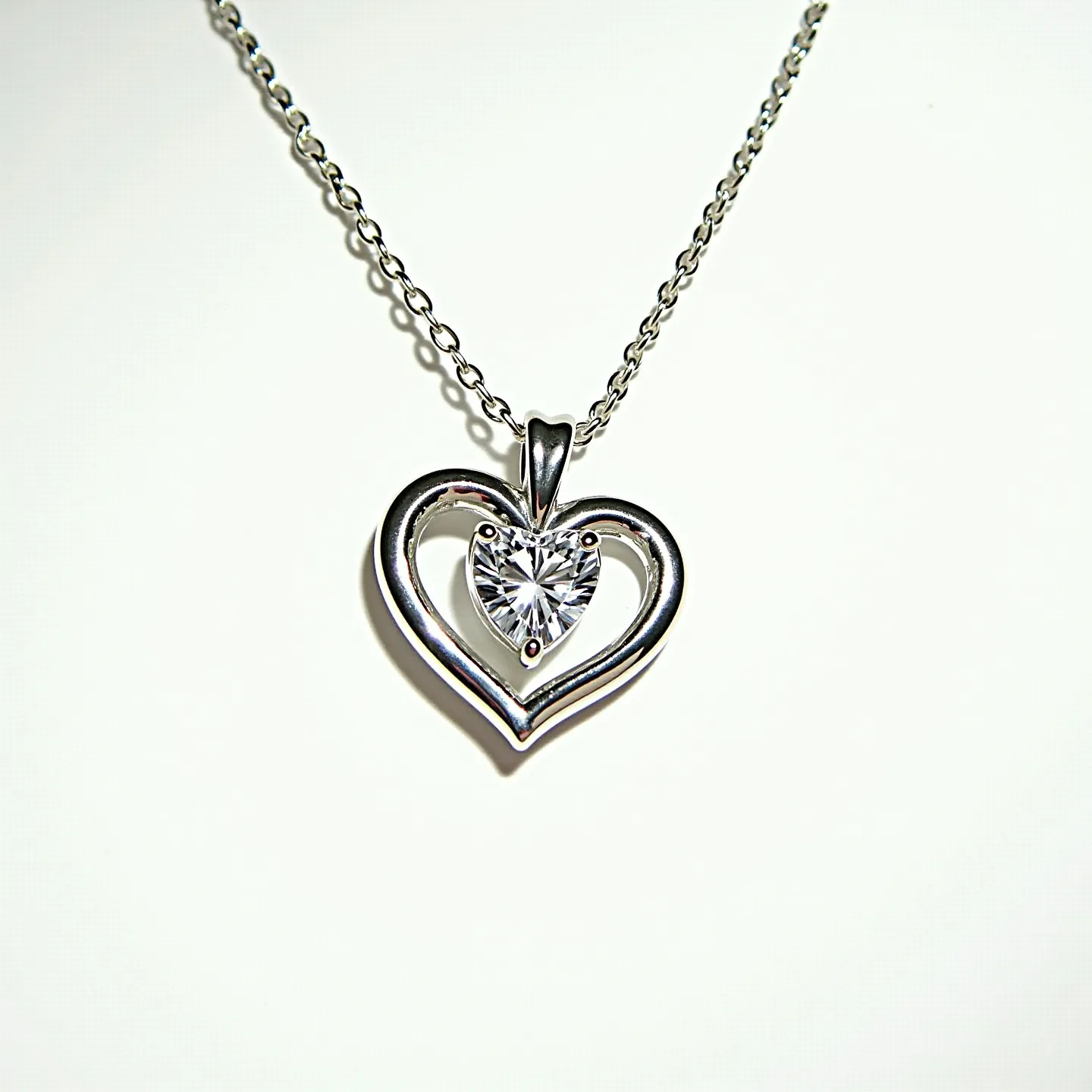 This diamond heart necklace features a delicate chain likely made of a shiny metal such as silver or white gold. The pendant consists of an elegant, open heart-shaped frame housing a central heart-cut gemstone, presumably a diamond, set in a prong setting that enhances its brilliance. The gemstone exhibits a clear, dazzling appearance, complemented by the polished finish of the surrounding metal. The necklace is equipped with a simple and secure clasp, ensuring ease of wear and stylish presentation.