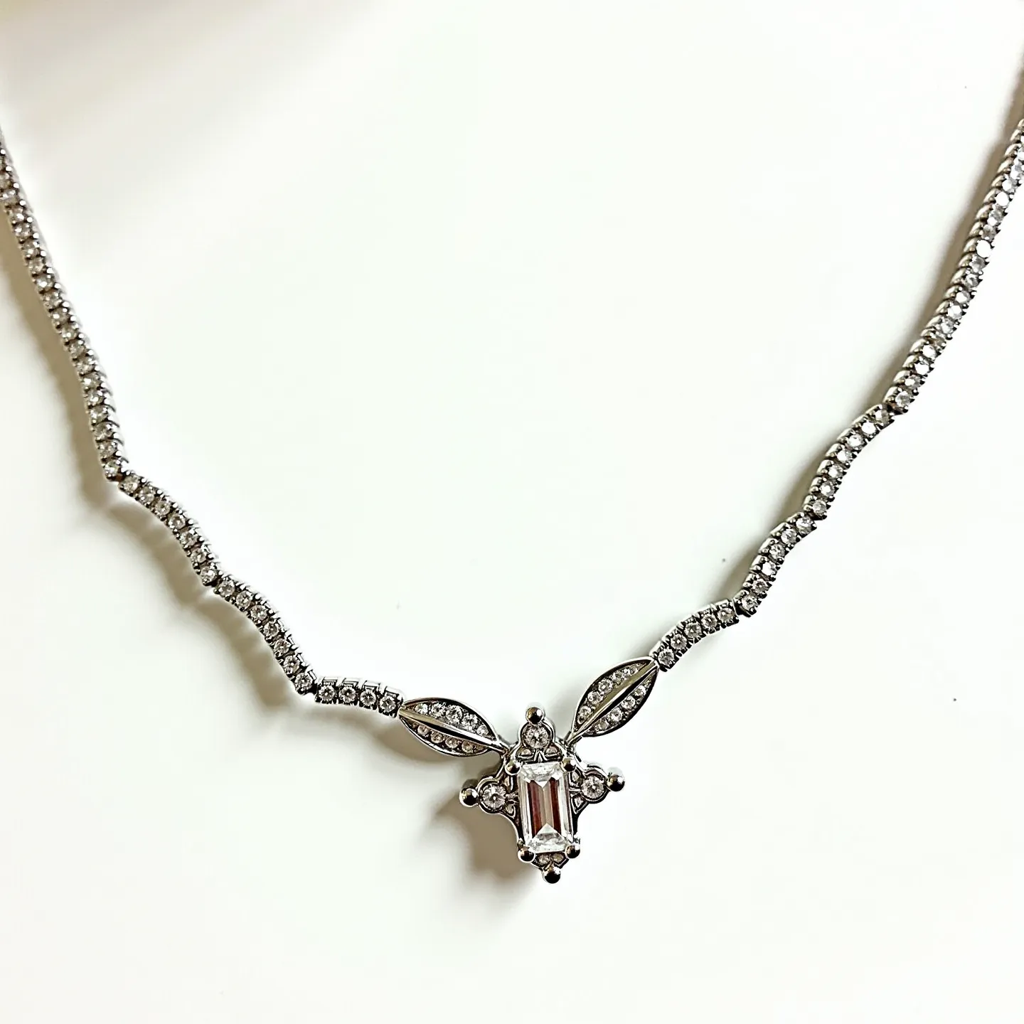 This diamond tennis necklace features a series of round-cut diamonds meticulously set along a sleek metal chain, likely white gold or platinum, which enhances their brilliance. The centerpiece of the necklace is an elaborate pendant with a central baguette-cut diamond surrounded by smaller round diamonds, set in an intricate design that includes marquise-shaped elements. The stones are held in secure prong settings, which allow for maximum light exposure and sparkle. The necklace is completed with a clasp that blends seamlessly with the overall design, ensuring both security and elegance in wear.