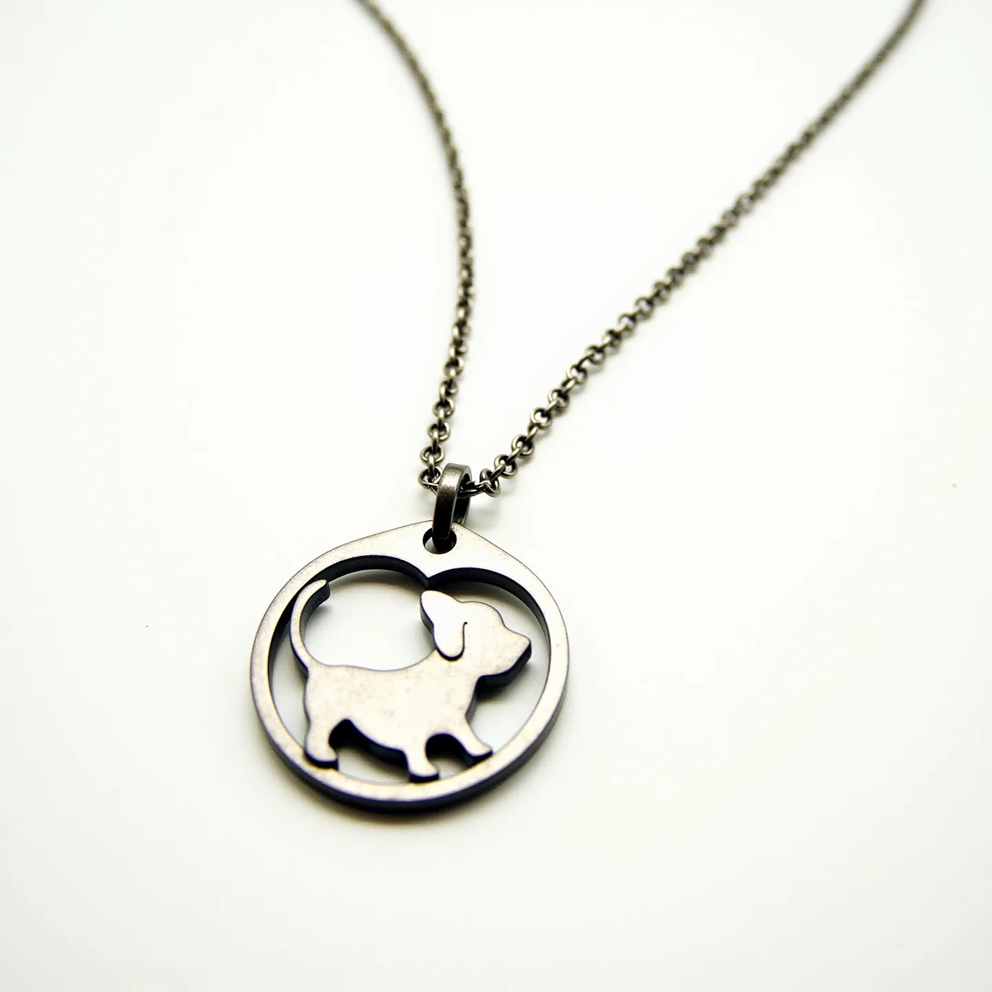 This dog necklace features a circular pendant made of metal with the silhouette of a dog design cut out in the center. The metal appears to be stainless steel, giving it a sleek and polished appearance. There are no gems or stones embedded in the pendant. It is attached to a simple metal chain via a small metal loop that acts as the clasp or connector, ensuring it hangs neatly. The chain is composed of small, interlinked loops that provide a smooth, continuous flow to the design.