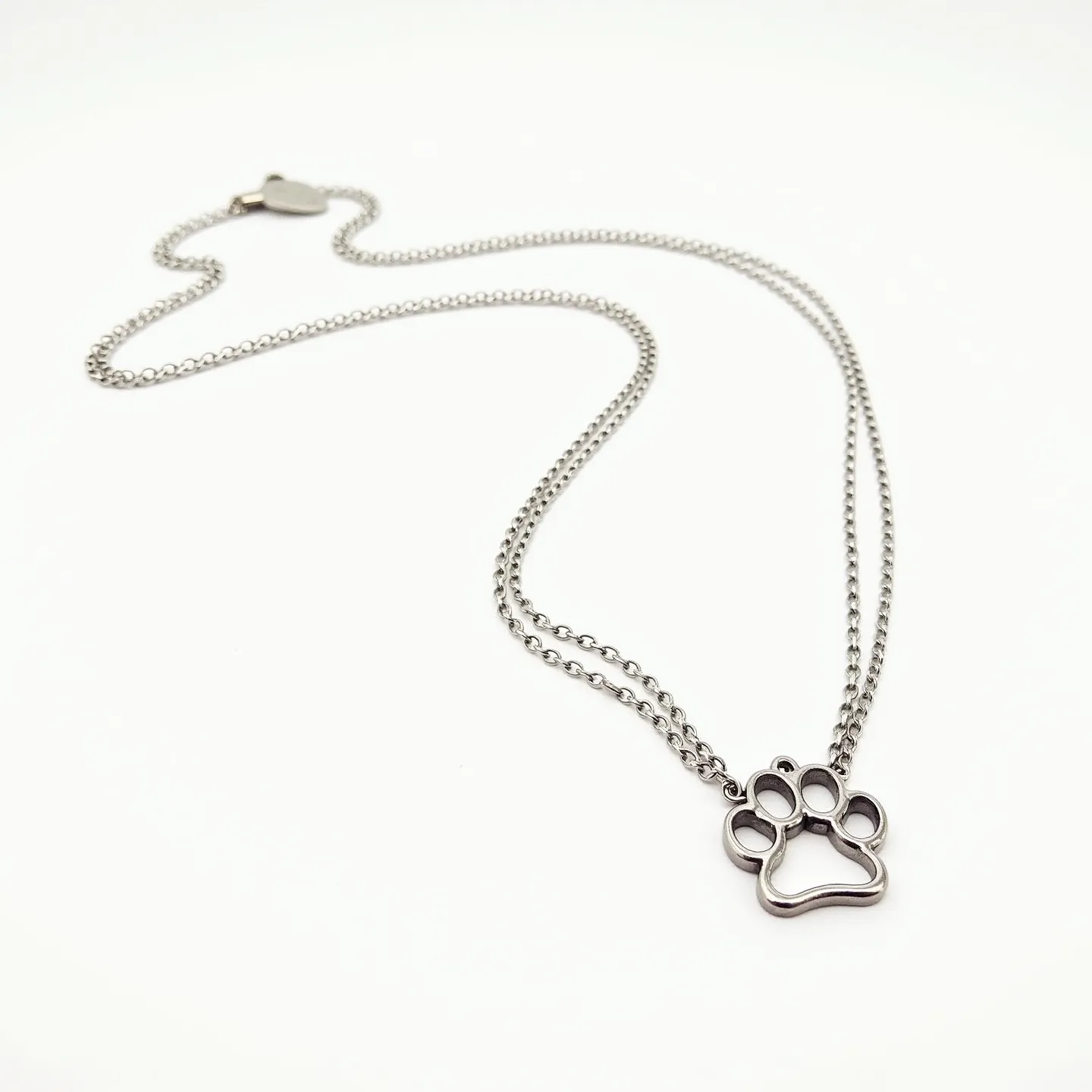 This dog necklace features a sleek and modern design with a metal chain and a pendant shaped like a dog paw. The pendant is crafted from a polished metal, possibly stainless steel, giving it a shiny and durable appearance. The chain is composed of interconnected links, providing both flexibility and strength. At the end of the chain lies a heart-shaped clasp, which allows for easy fastening and secure wearing. The simple yet stylish design makes it a versatile accessory for any dog lover. No gemstones or intricate settings are present, enhancing its minimalist charm.