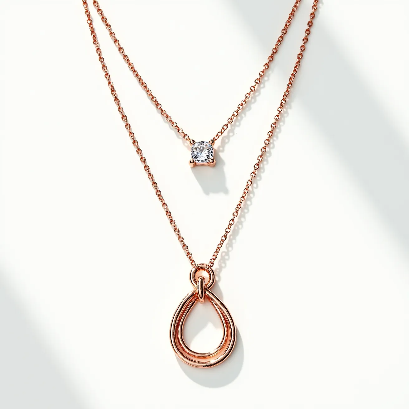 This double necklace features two delicate, rose gold-toned chains of varying lengths. The shorter chain includes a single, round-cut gem, likely a diamond or a cubic zirconia, set in a four-prong setting that allows maximum light reflection. The longer chain showcases a smooth, teardrop-shaped pendant, elegantly simple in its design with a polished finish that complements the overall aesthetic. Both chains are likely connected by a discreet clasp at the back, maintaining the necklace's minimalist and refined style.