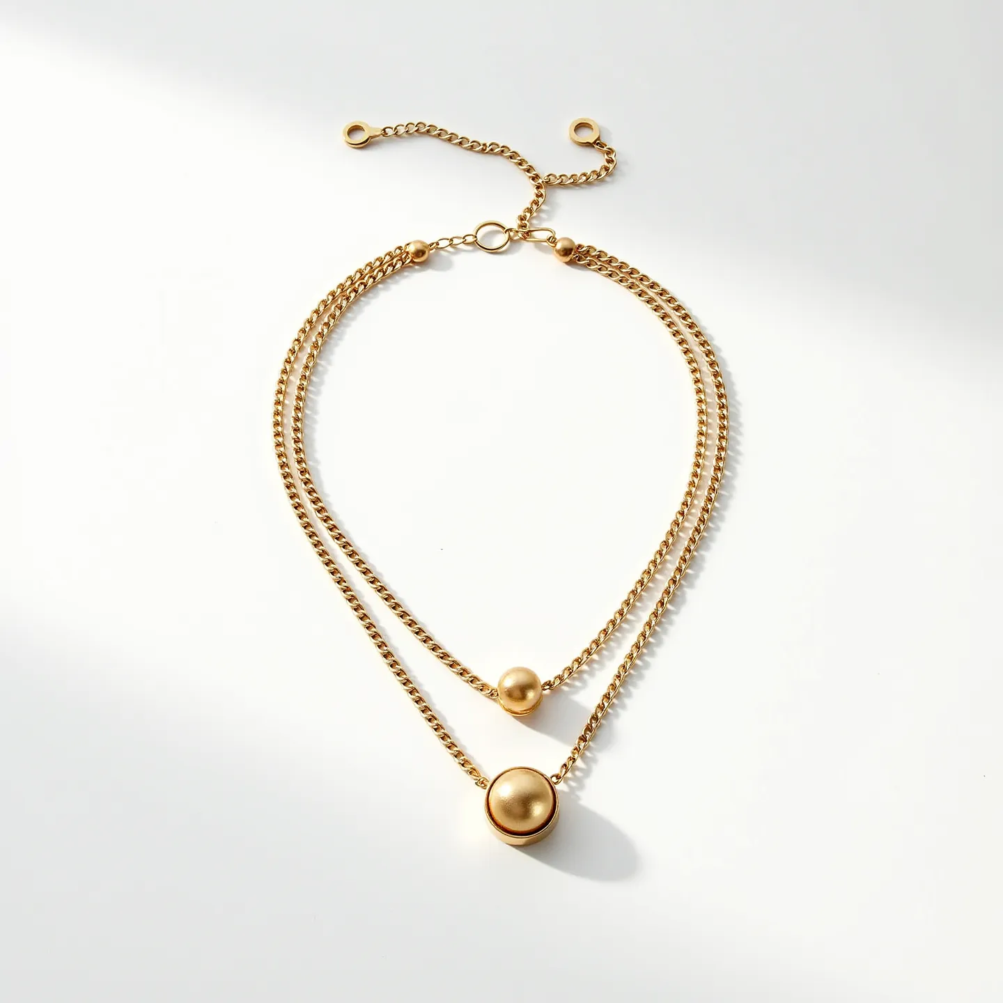 This double necklace features two strands of gold-toned chains, with each strand hosting a single spherical pendant. The upper strand has a smaller gold-colored sphere, while the lower strand showcases a larger similar sphere, each resting elegantly on the chain. Both spheres appear smooth and polished, capturing light softly. The necklace is completed with an adjustable clasp system, allowing for varying lengths to suit the wearer’s preference. The overall design is sleek and minimalistic, offering a classic, sophisticated touch to any ensemble.