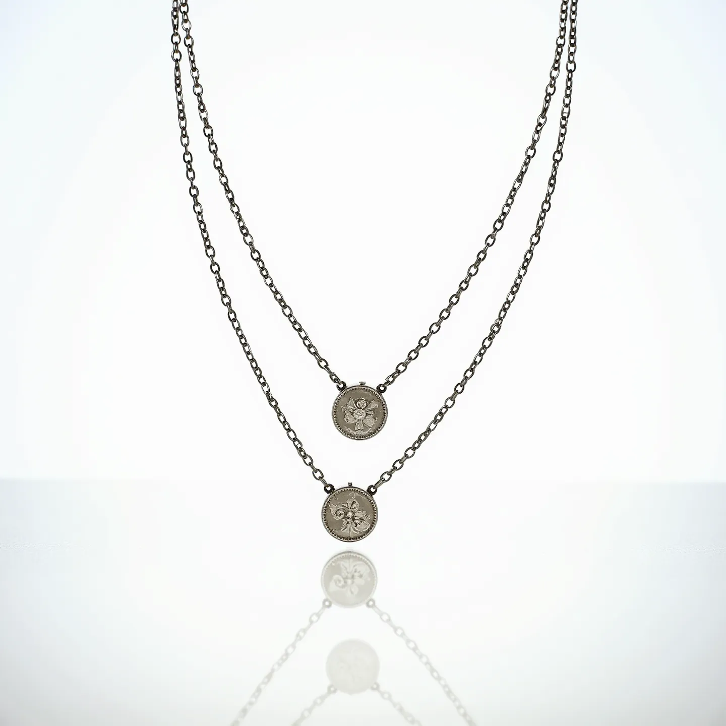 This double necklace features two delicate chains, likely crafted from a silvery metal, each suspending a disc-shaped pendant with intricate designs. The pendants are circular and have engraved patterns; the top pendant displays a floral motif, while the lower one features a fleur-de-lis design. Both pendants are framed by a subtle beaded edge, enhancing their visual appeal. The chains are made of simple, evenly sized links that complement the understated elegance of the pendants. The necklace likely fastens with a standard clasp, ensuring a secure fit while allowing the pendants to rest comfortably at different lengths for a layered look.