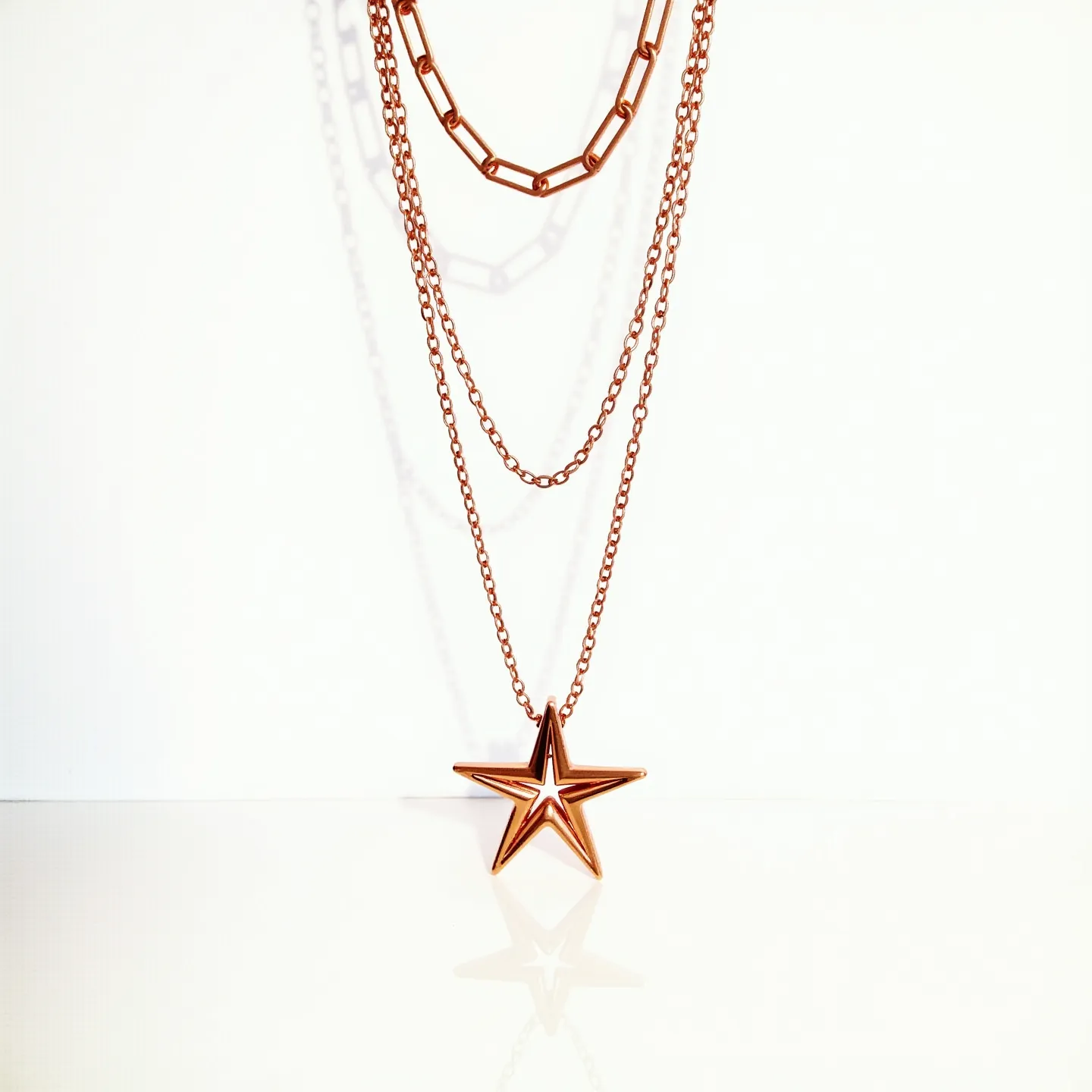 This double necklace features a combination of chain styles, including a bold, chunky link chain paired with delicate, slender chains. It is crafted from a polished metal with a warm, rose-gold hue that adds a touch of elegance. The necklace is adorned with a striking star-shaped pendant, which is open and geometric, contributing a modern design element. The construction suggests a seamless integration with no visible clasp, allowing the necklace to drape gracefully. The materials and design create a harmonious balance of contemporary chic and sophistication.