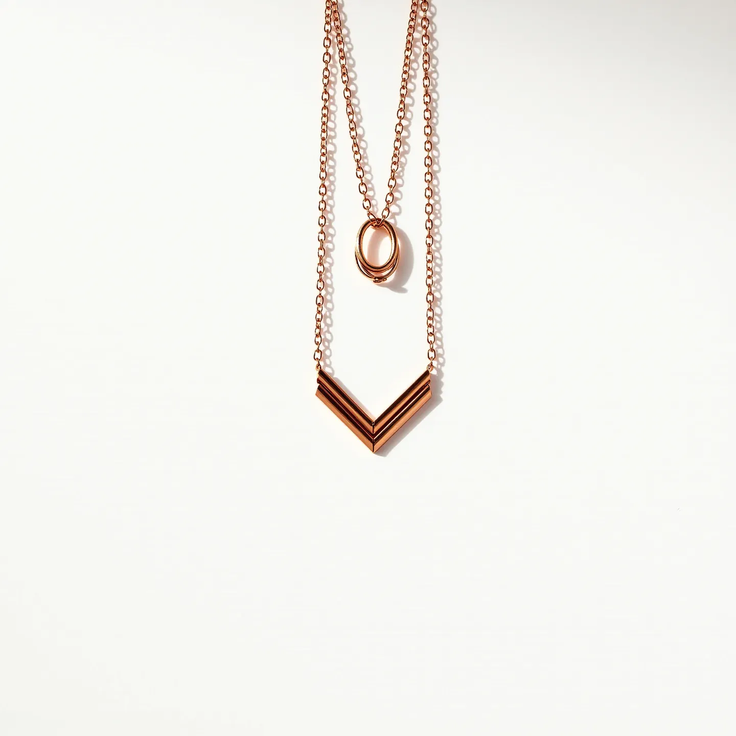 This double necklace features two distinct chain lengths made from a polished metal, which appears to be rose gold in color. The shorter chain is adorned with an intertwined circular pendant, showcasing a sleek, minimalist design. The longer chain holds a V-shaped pendant comprising narrow, parallel bars that add a geometric element to the piece. The clasps connecting the necklace chains are not visible in the provided image. This elegant design highlights a contemporary aesthetic without the inclusion of any gemstones.