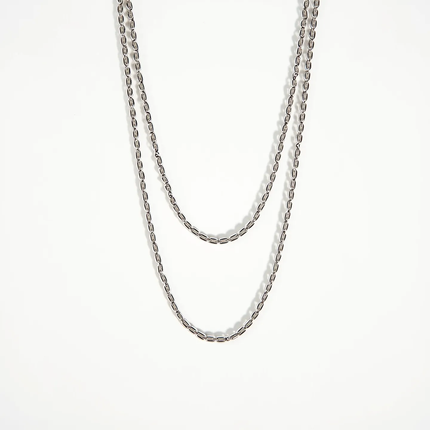 This double necklace features a sleek and modern design with two chains made of metal links, likely crafted from stainless steel or a similar shiny silver-toned material. The chains are uniform in size and texture, featuring elongated, oval links that create a fluid and elegant silhouette. Each chain is of a different length, allowing for a layered look when worn. The necklace likely incorporates a clasp for fastening, possibly a lobster or spring ring clasp, which ensures secure wear. There are no gemstones or additional embellishments on the chains, highlighting the minimalist aesthetic of the piece.