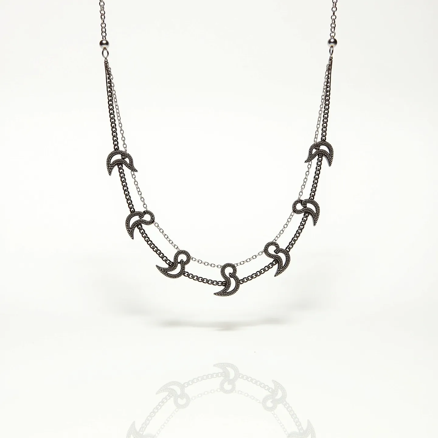 This double necklace features an elegant design with two parallel metal chains. Each chain is adorned with smooth, curved crescent-shaped pendants that interconnect gracefully. The crescents are meticulously detailed with a textured surface, possibly made from a polished metal that enhances their visual appeal. The necklace includes a small bead-like element near the clasp area, adding a subtle decorative touch. The chains connect at each end with a traditional clasp, ensuring secure and adjustable fastening. The overall design is both modern and versatile, suitable for various occasions.