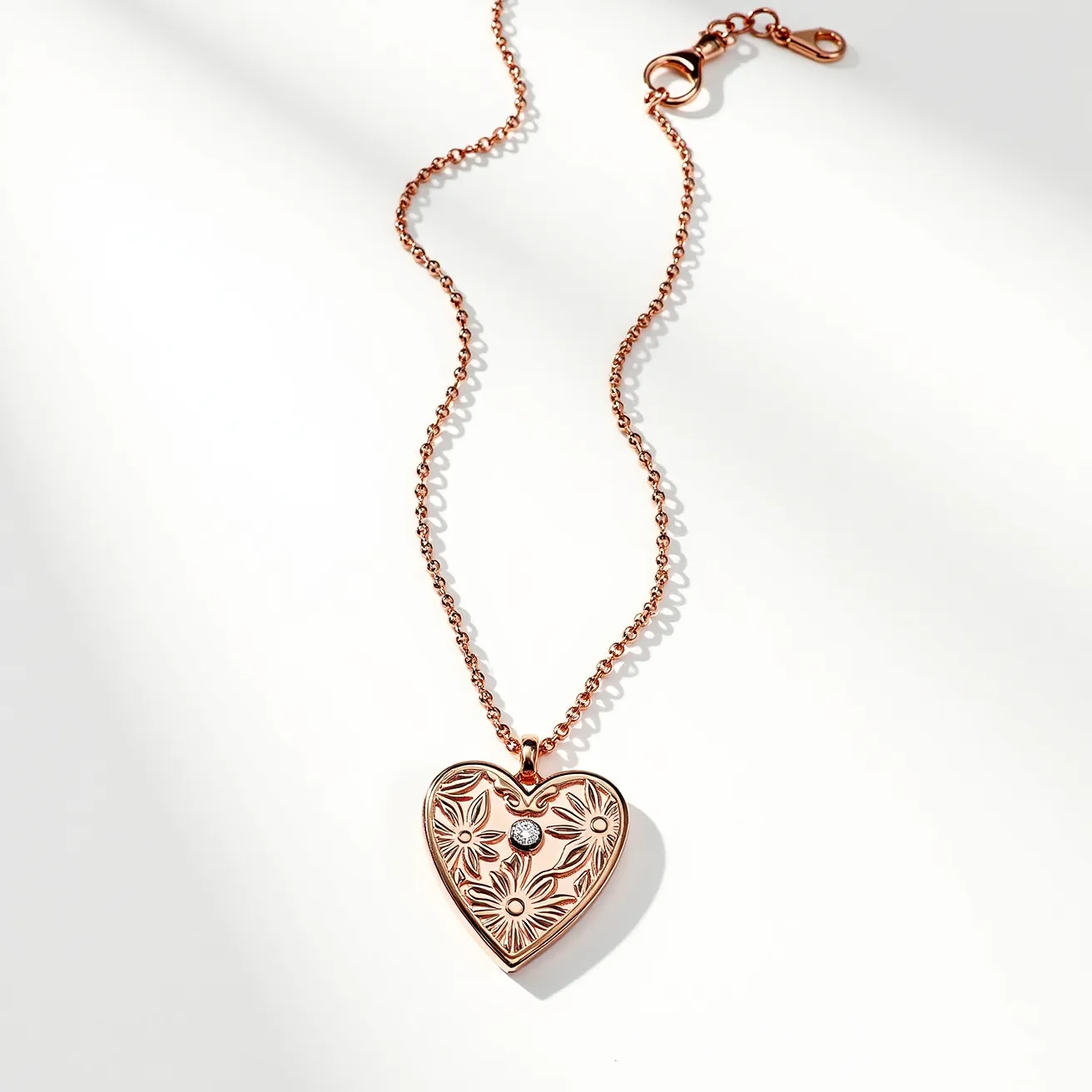 This engraved heart necklace features a rose gold-toned metal chain and pendant, showcasing a detailed floral design. At the center of the heart-shaped pendant, there is a single sparkling gemstone, likely a round-cut diamond or cubic zirconia, set securely in a bezel setting to add an elegant touch. The chain is composed of small, evenly linked loops, culminating in a lobster clasp and an extender chain that allows for adjustable length, providing both functionality and style. The necklace embodies a blend of romantic aesthetics and practical craftsmanship.