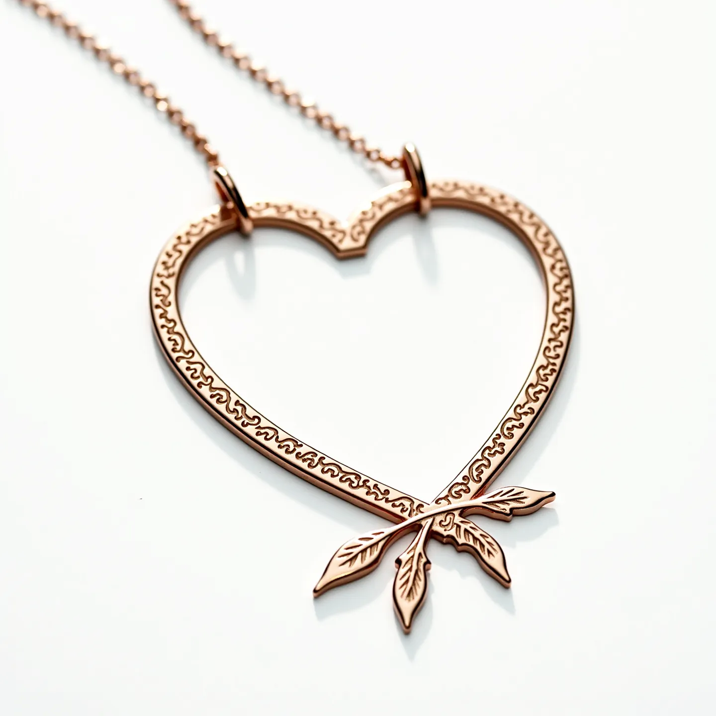 This engraved heart necklace is crafted from a polished metal with a warm, rose gold hue. The heart outline features intricate engraving, which adds a touch of elegance to its design. At the bottom, two detailed leaves are attached, complementing the heart's border. It is suspended by a delicate chain, attached through two circular loops on the upper sides of the heart, ensuring it lays centered. The necklace design is both graceful and sophisticated, ideal for adding a subtle statement to any outfit.