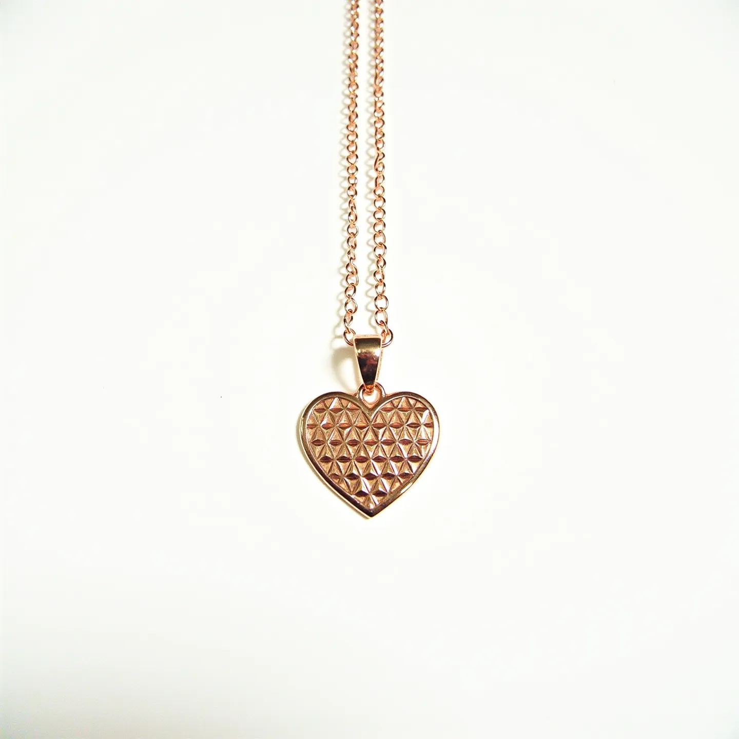 This engraved heart necklace features a rose gold-tone metal heart pendant with an embossed geometric pattern on its surface, creating a textured and reflective finish. The pendant is elegantly suspended from a matching rose gold-tone chain, ensuring a cohesive design. The pendant is connected to the chain via a rose gold-tone bail, adding a sleek transition between the chain and the heart. The chain likely includes a traditional clasp mechanism, such as a lobster claw or spring ring clasp, for secure attachment and ease of wear.