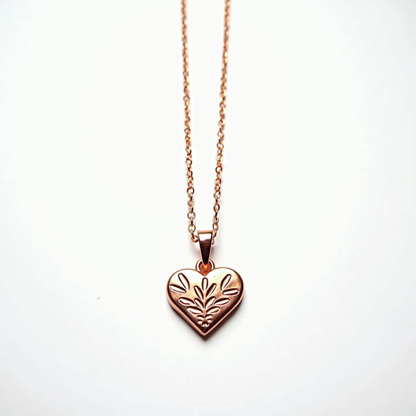 This engraved heart necklace features a delicate heart-shaped pendant crafted from a warm-toned metal, likely rose gold or copper. The pendant is intricately designed with a floral pattern engraved into its surface, adding an elegant touch. The heart is suspended from a chain composed of small round links that match the pendant's metal. The necklace is secured with a standard lobster clasp, ensuring reliability and ease of use.