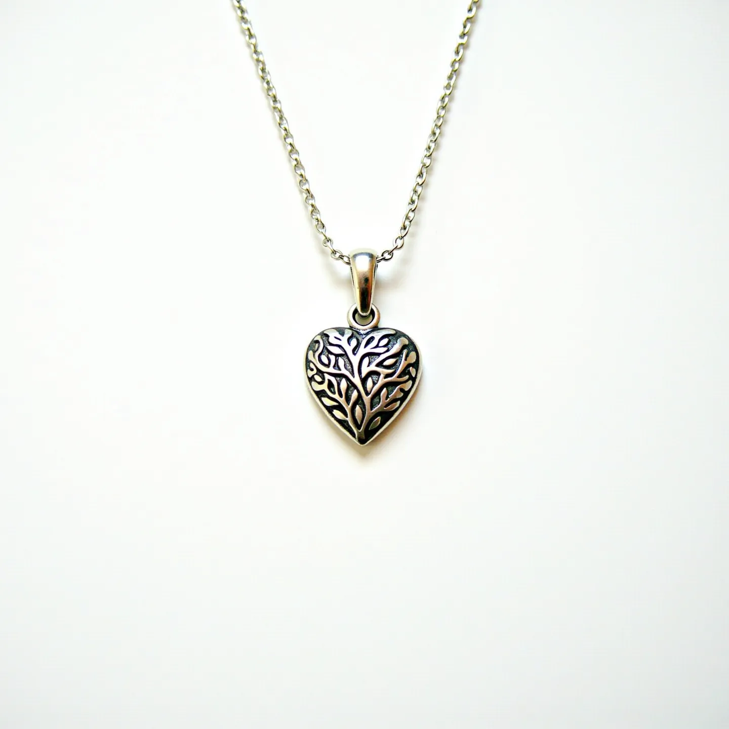 This engraved heart necklace features a beautifully detailed heart pendant made of shiny metal, possibly silver or stainless steel, showcasing an intricate tree of life design. The surface of the pendant displays a raised pattern, giving it a textured, elegant appearance. The pendant is attached to a delicate chain through a solid bail, which provides a steady and secure hold. The chain is composed of small, interlinked oval links, offering a classic and timeless aesthetic. The necklace likely uses a standard clasp, ensuring it is easy to put on and take off.