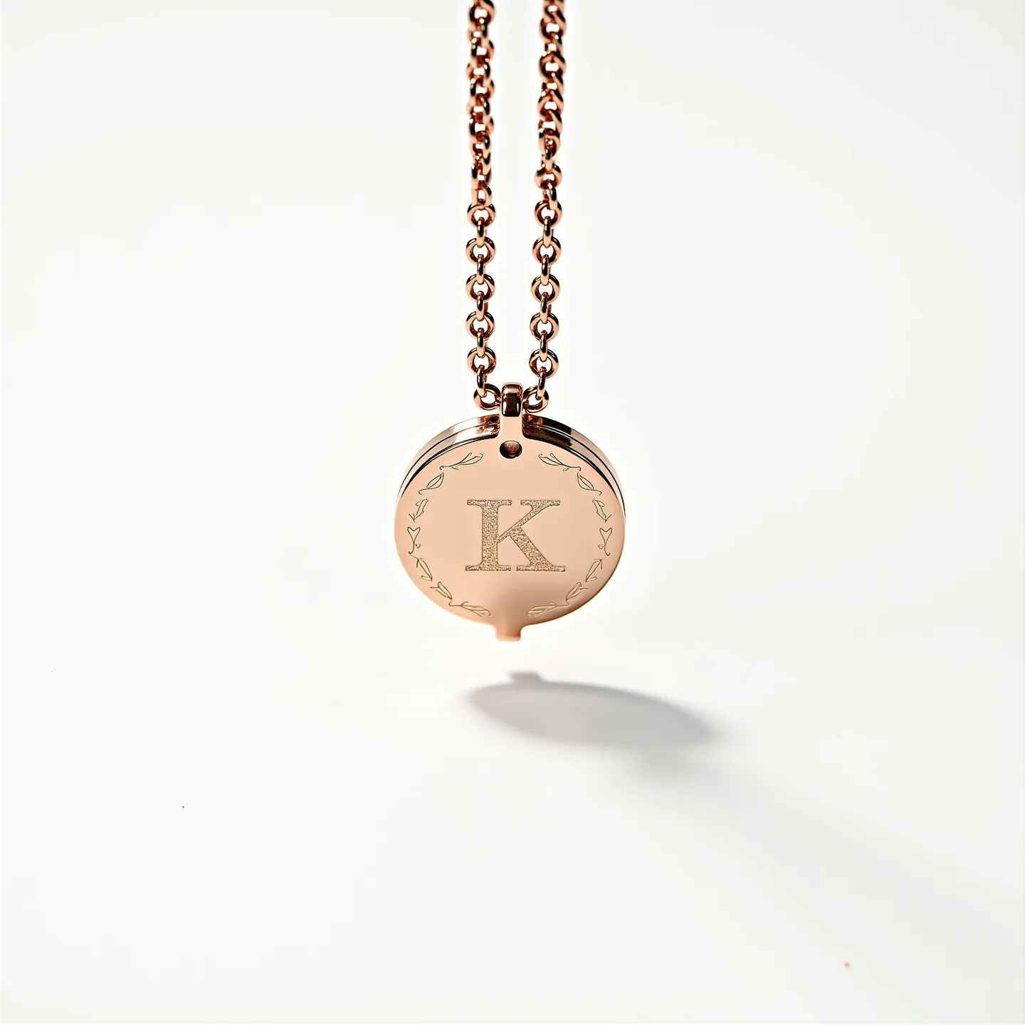 This engraved necklace features a circular pendant crafted from a metallic material, likely rose gold in appearance, hanging from a coordinating chain of interlinked round elements. The pendant is intricately engraved with the letter "K," surrounded by a decorative border of vine-like designs. The chain is attached to the pendant via a sturdy loop at the top, suggesting a simple yet secure method of connection. The overall design highlights elegance and personalization, making it a stylish accessory with a personalized touch.