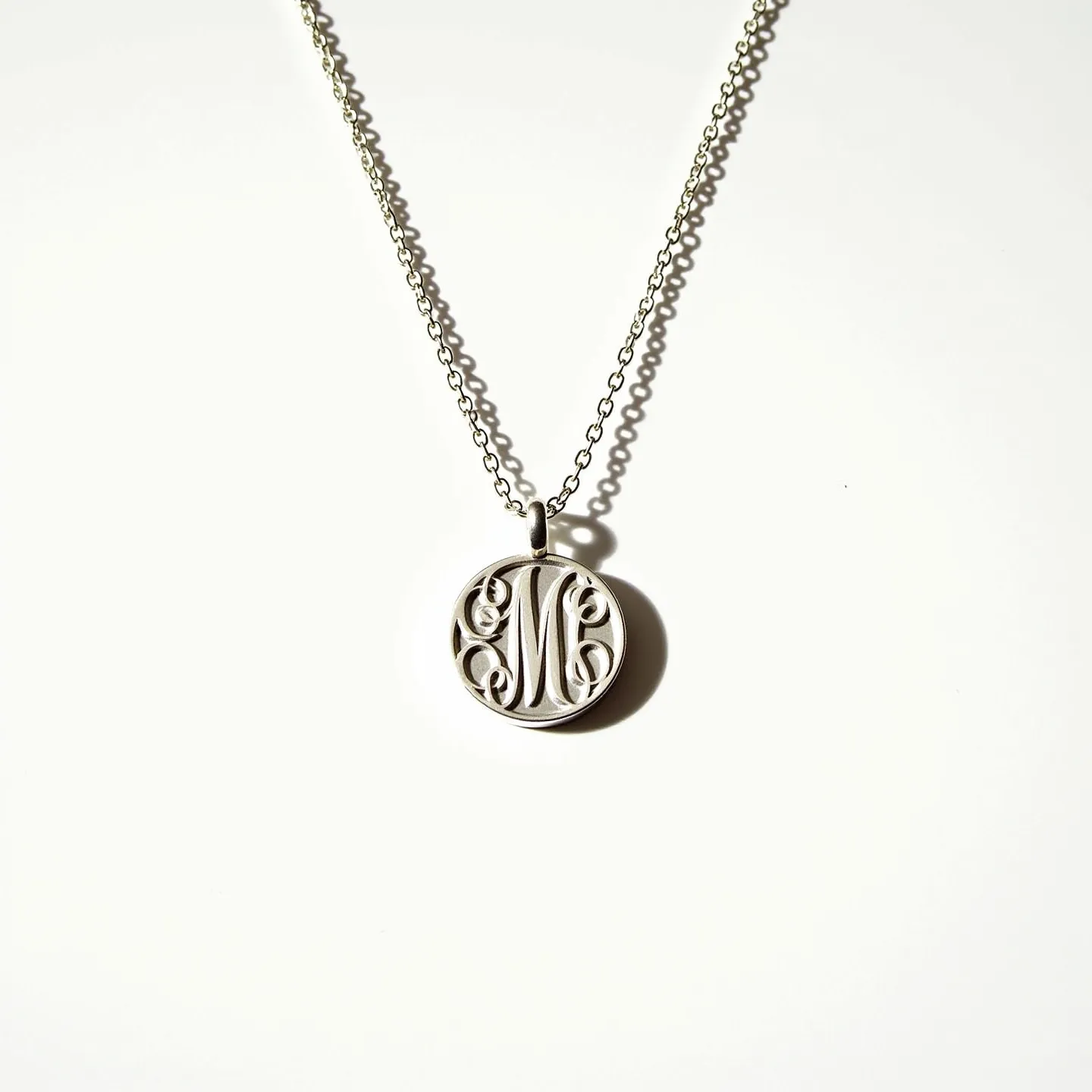 This engraved necklace features a circular pendant with a beautifully engraved letter "M" in an intricate, scrolling script. The pendant appears to be crafted from a silver-toned metal, giving it a classic and elegant look. It is suspended on a delicate, matching chain made of small, round links, ensuring it hangs elegantly around the neck. The necklace is fitted with a simple loop attachment, seamlessly integrating the pendant with the chain. This type of design highlights personal expression, while maintaining a refined and timeless appearance.