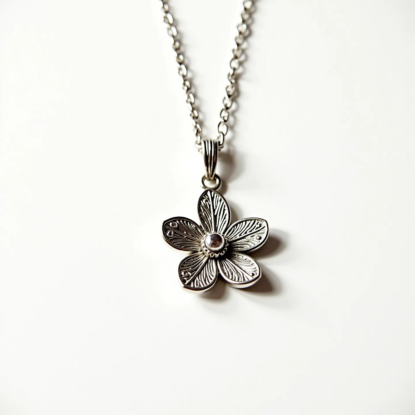 This flower necklace features a delicate flower pendant crafted from silver-toned metal with intricately engraved petals, giving it a lifelike appearance. At the center of the flower is a small, round gemstone, possibly a clear quartz or diamond, set in a simple bezel setting that securely positions the gem while highlighting its clarity. The necklace is attached to a sturdy chain made of similar silver-toned metal, ensuring durability and a cohesive look. The pendant is connected to the chain by a decorative bail that adds an ornamental touch to the piece. The chain is linked by an easy-to-use clasp that ensures the necklace is both secure and comfortable to wear.