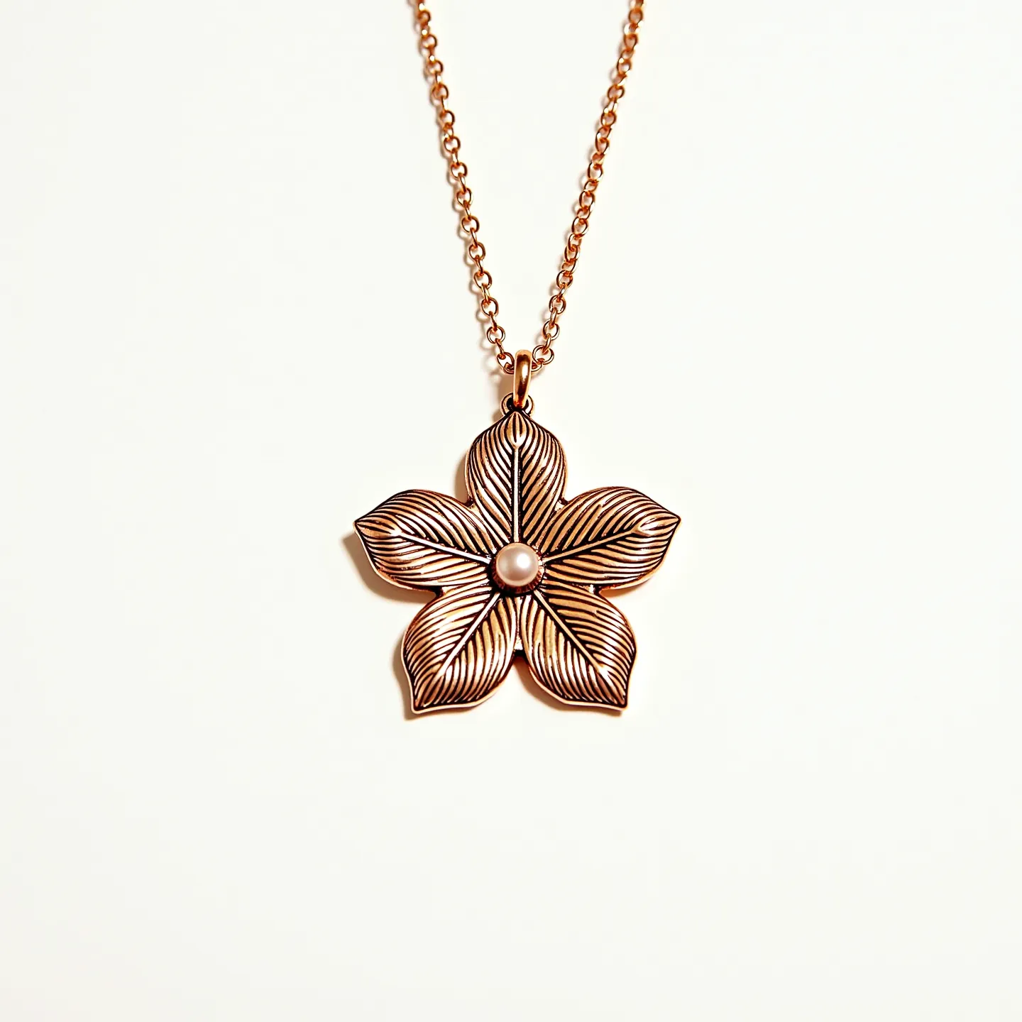 This flower necklace features a beautifully crafted floral pendant made of metal with an intricate pattern that gives the petals a textured appearance. At the center of the flower is a round, polished pearl set smoothly into the metal, adding a touch of elegance. The pendant is suspended from a chain with a warm, rose gold tone, which complements the metalwork of the flower. The necklace is equipped with a standard chain clasp, ensuring it is secure and easy to wear.